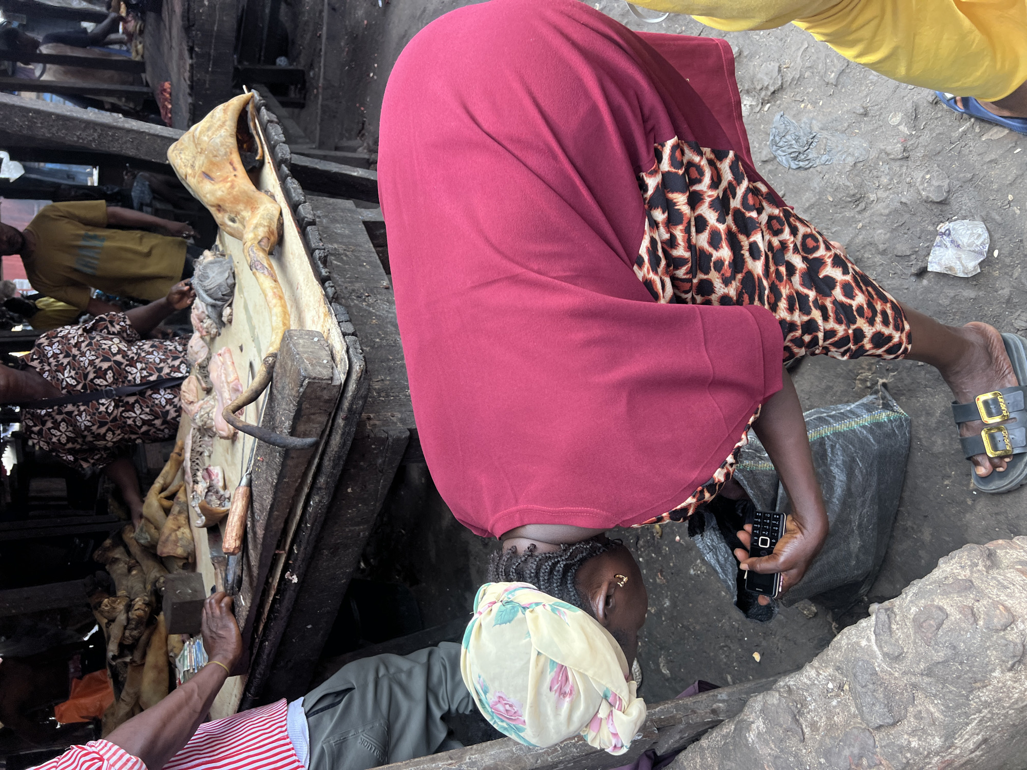 In Nkpor, Nigeria, a scene unfolds at what appears to be a bustling open-air market. The central figure, a woman wearing a vibrant red dress and a floral headscarf, is bent over, examining a black mobile phone in her hand. Her patterned skirt, adorned with a leopard print, peeks out from beneath the red fabric. Beside her, a dark plastic bag lies on the ground, and a stone obstacle is visible to her left.
The market environment is characterized by wooden stalls laden with various items, likely meat products, as indicated by the raw cuts and animal parts displayed on a long table in the background. A man in a red and white striped shirt stands to the left, his arm resting on the table, while another man in a yellow t-shirt is partially visible further back. The ground is rough and appears to be dirt or packed earth, with scattered debris. The overall impression is one of a busy commercial hub, with a focus on fresh produce and lively community interaction.