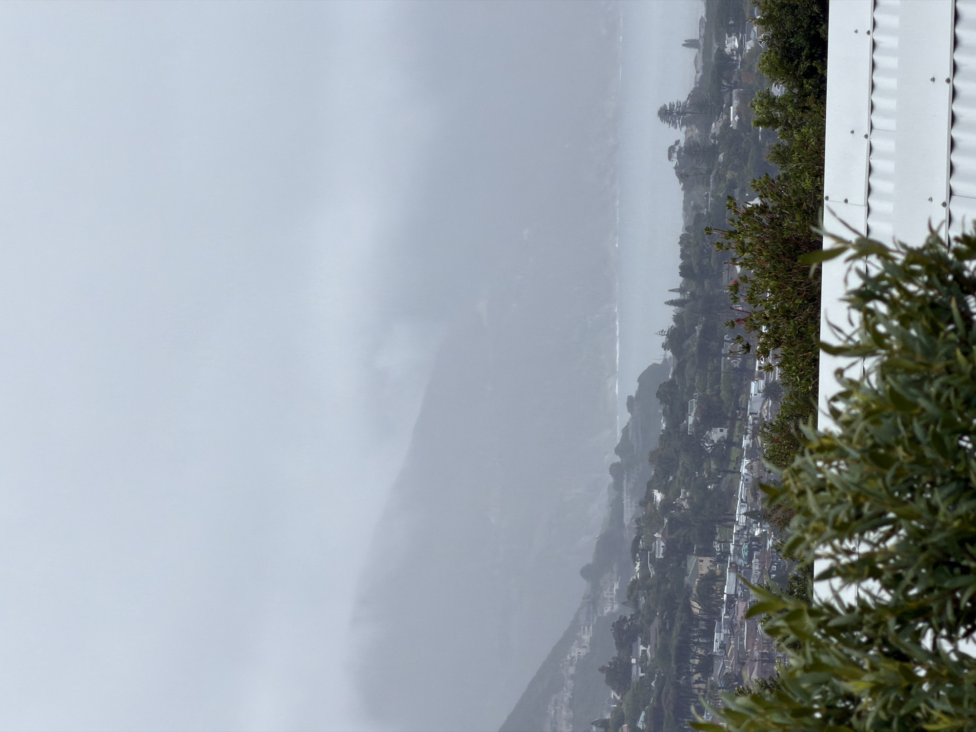 The image shows a misty, overcast view of Hout Bay, South Africa. The left side of the image is dominated by a fog-shrouded mountain. The ocean is visible as a sliver in the middle ground. A hillside dotted with houses and trees slopes towards the water. In the foreground, a white corrugated wall and some green foliage are partially visible, framing the scene.