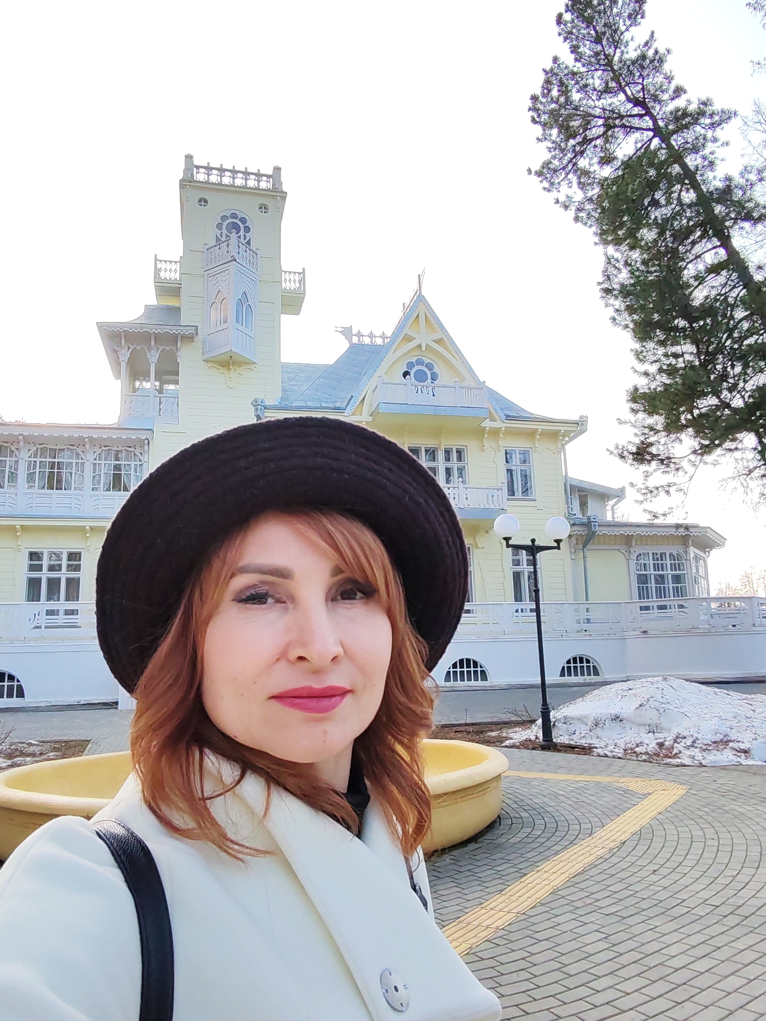 A selfie features a woman with auburn hair wearing a black hat and a white coat with a black shoulder bag. She is positioned in front of a yellow building featuring ornate architectural details, including towers, balconies, and windows. A tall tree stands to the right of the building, extending into the upper right corner of the frame.
The woman is situated on a paved area with a yellow planter visible in the foreground. Patches of snow are present behind the planter. A lamppost stands between the woman and the building.
The location is identified as Sarapul, Russia.