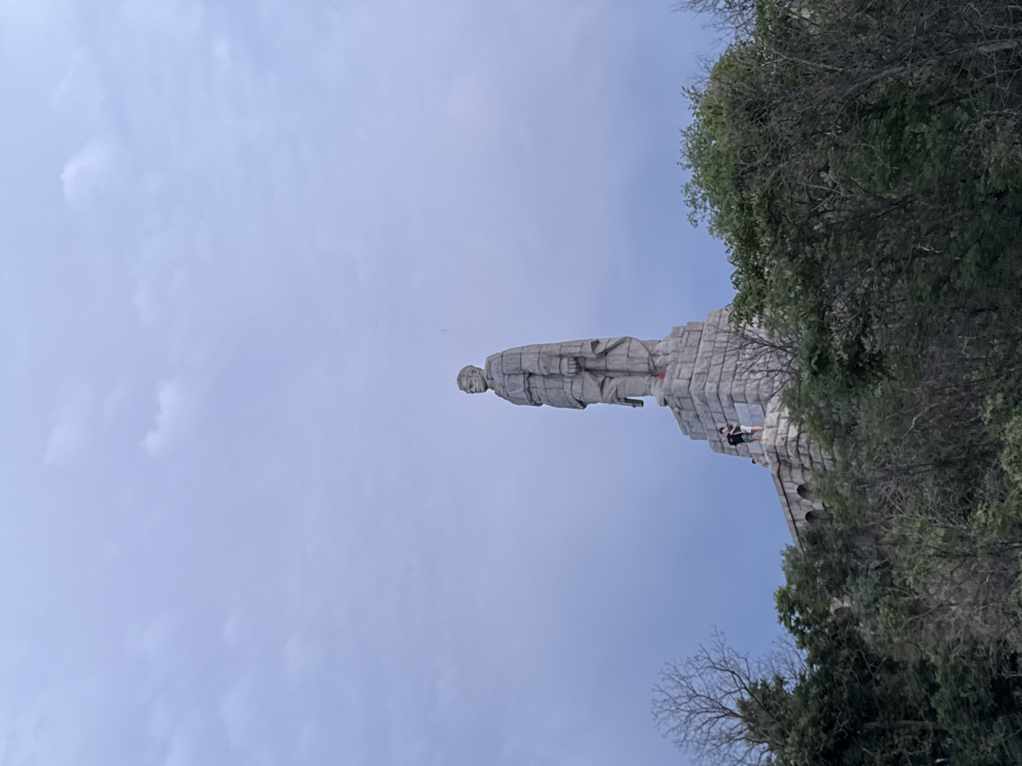 The image displays the towering Alyosha Monument in Plovdiv, Bulgaria, featuring a large grey stone statue of a male figure, identified as a Soviet soldier. The statue stands prominently on a massive, tiered stone pedestal, depicted from a low-angle perspective that emphasizes its height. The soldier is clad in a military greatcoat, holding what appears to be a rifle pointed downwards.
The monument is set against a bright, light blue sky with scattered faint white clouds, indicating daytime and fair weather. To the lower right of the statue, on a section of the pedestal, two small human figures are visible. These individuals appear to be standing or walking on the monument's structure, providing a sense of scale to the colossal statue and suggesting a visit to the site. The right side and foreground of the image are partially framed by green foliage from trees and some bare tree branches, implying a park-like or natural setting surrounding the monument. No discernible text is visible on the monument or in the immediate surroundings.