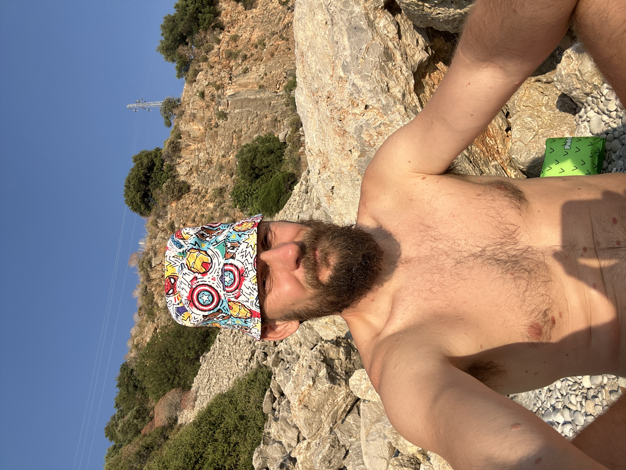 An outdoor, eye-level selfie shows an adult male seated on a rocky and pebbled shoreline, consistent with the coastal environment of Fethiye, Turkey. The man, featuring a dark brown beard and bare chest, wears a brightly colored bucket hat adorned with various Marvel Comics character logos, including Captain America's shield, Spider-Man's symbol, and Thor's hammer. He is looking directly at the camera with a slight smile. His skin is tanned and displays scattered red dermal markings on his chest and arms. To his lower left, partially obscured, a green object with the visible text "Pak" and a pattern of white "V" shapes rests on the pebbles. The background features a steep, arid hillside composed of reddish-brown rock formations and sparse green vegetation. High on the hill, a metal lattice electrical transmission tower and associated power lines are visible against a clear, deep blue sky. Strong sunlight illuminates the scene, casting distinct shadows.