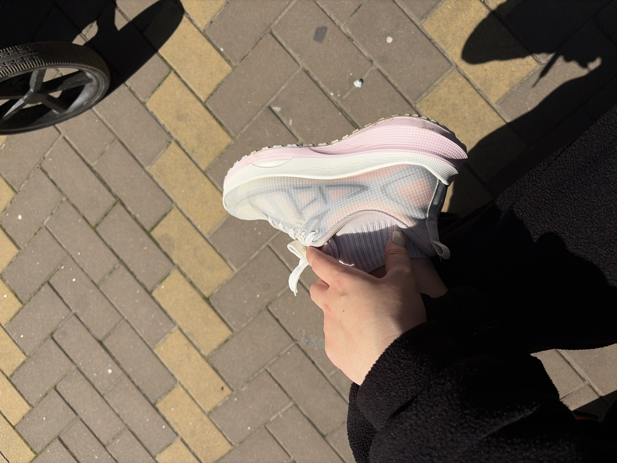 The image shows a person holding a light pink and white running shoe on a brick sidewalk. The sidewalk is made of bricks in a herringbone pattern, with alternating rows of tan and grey bricks. The person is wearing a black coat or sweater. In the upper left corner of the image is a tire or wheel. The setting is outdoor, likely in a public space. The city of Krasnodar, Russia, cannot be determined from this image.