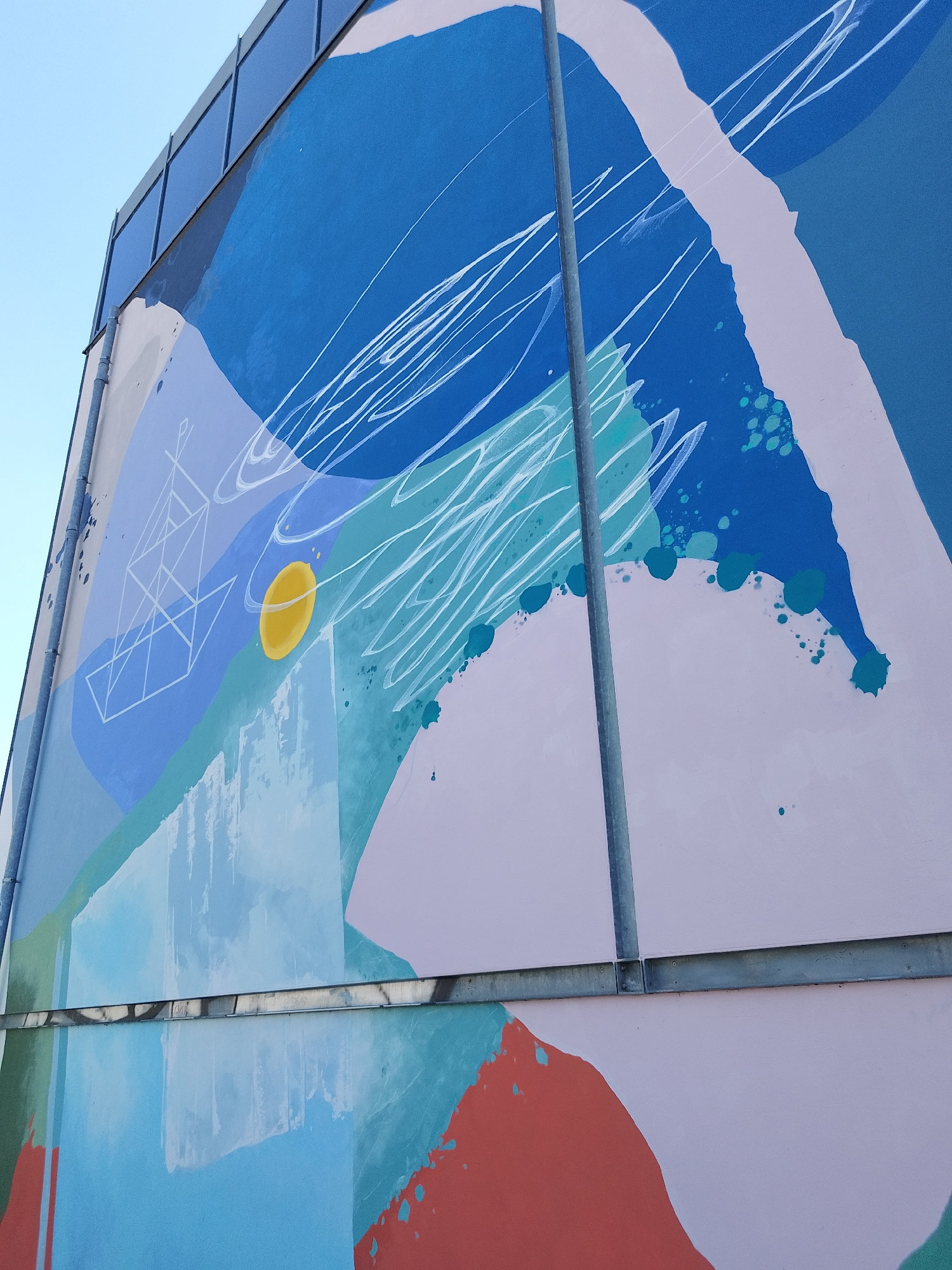 The image shows a segment of a building exterior featuring a large abstract mural. The mural is composed of various shapes and colors including blues, greens, lavender, and red-orange. There are also elements resembling white lines or scribbles, and geometric figures. The background sky is a clear blue. The location is Unknown, Unknown.
