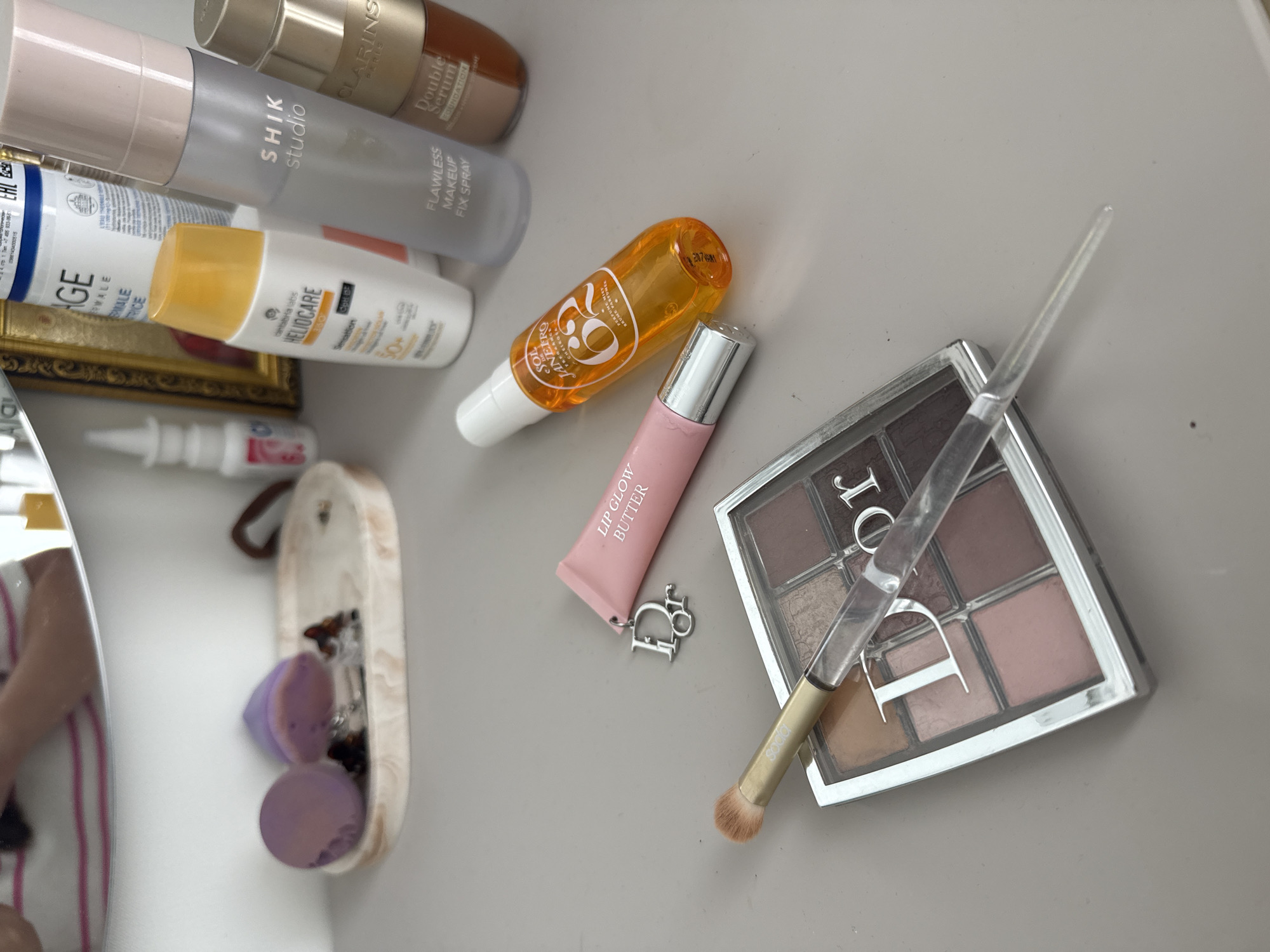 This flat lay image captures a collection of beauty and skincare products arranged on a light gray surface, likely a vanity or dressing table in Murom, Russia. A partial reflection of a person wearing a striped garment is visible in a mirror on the left.
The array of objects includes several bottles and tubes of cosmetic and skincare items: a "CLARINS PARIS Double Serum FOUNDATION," a "SHIK studio FLAWLESS MAKEUP FIX SPRAY," a "SOL JANERRO CHEERGE 62 PERFUME MIST," and a "cantabria labs HELIOCARE 360" sunscreen with "50+" SPF. Other partially visible texts on bottles include "AGE," "EPWALE," "MALE," and "RICE." A pink tube labeled "LIP GLOW BUTTER" is also present.
Next to the lip product, a silver-toned D-shaped charm, likely from Dior, is visible. Adjacent to these items is an open makeup palette, predominantly featuring brown and pink eyeshadow shades, with the letters "IO R" prominently displayed as part of its design, suggesting it is a Dior product. A makeup brush with a golden handle, bearing the brand "soda," rests on the palette.
Further to the left, a wooden holder contains two purple makeup sponges. The lighting appears bright and even, suggesting daytime. The overall arrangement implies a personal beauty routine setting.