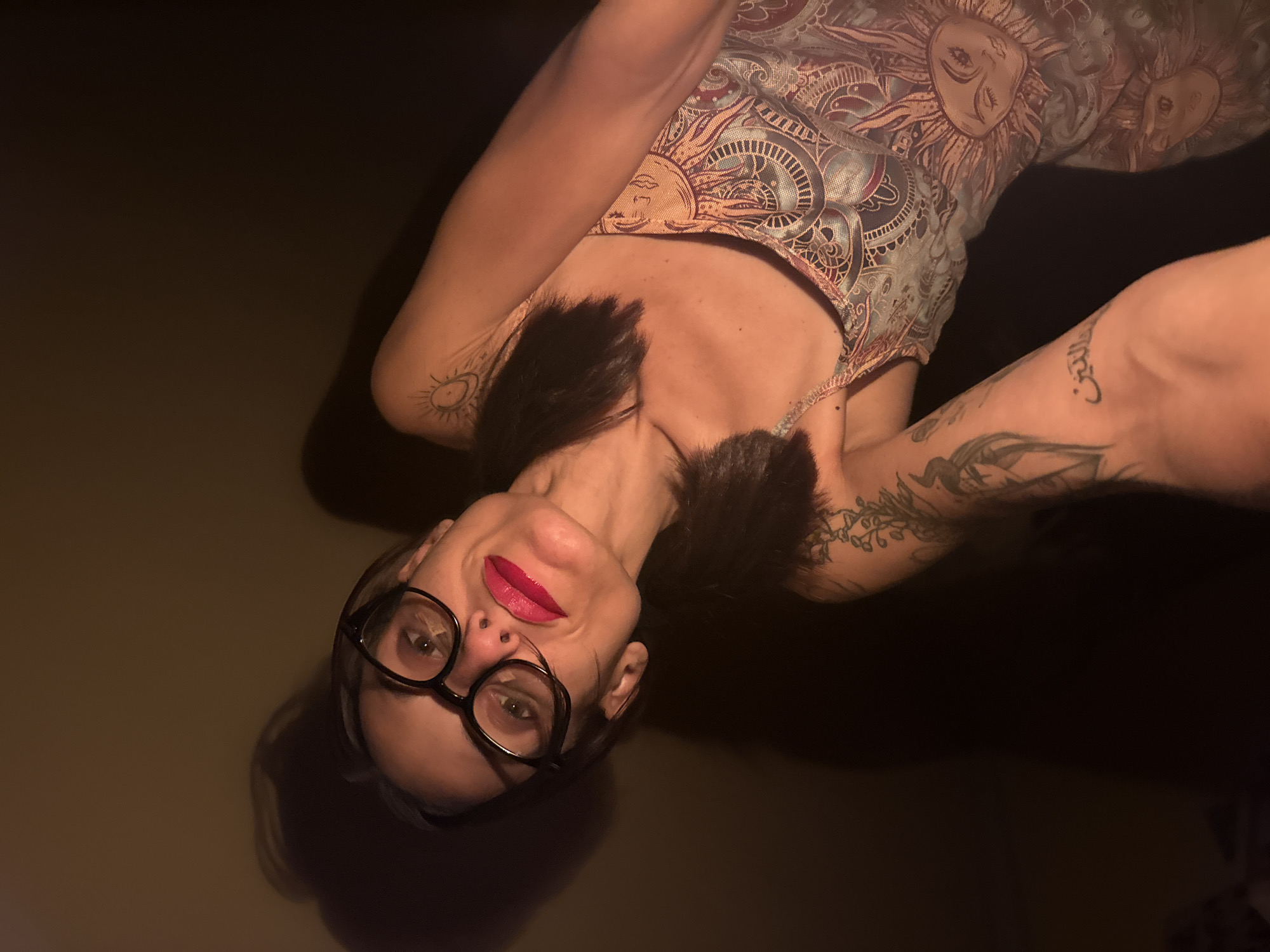 A woman with dark hair and dark-rimmed glasses, wearing bright red lipstick, poses for a selfie. She is wearing a patterned tank top with celestial motifs. Her left arm, visible in the foreground, has tattoos. The background is dark and indistinct, suggesting an indoor setting with dim lighting. There is no discernible information about the location in Pleven, Bulgaria.