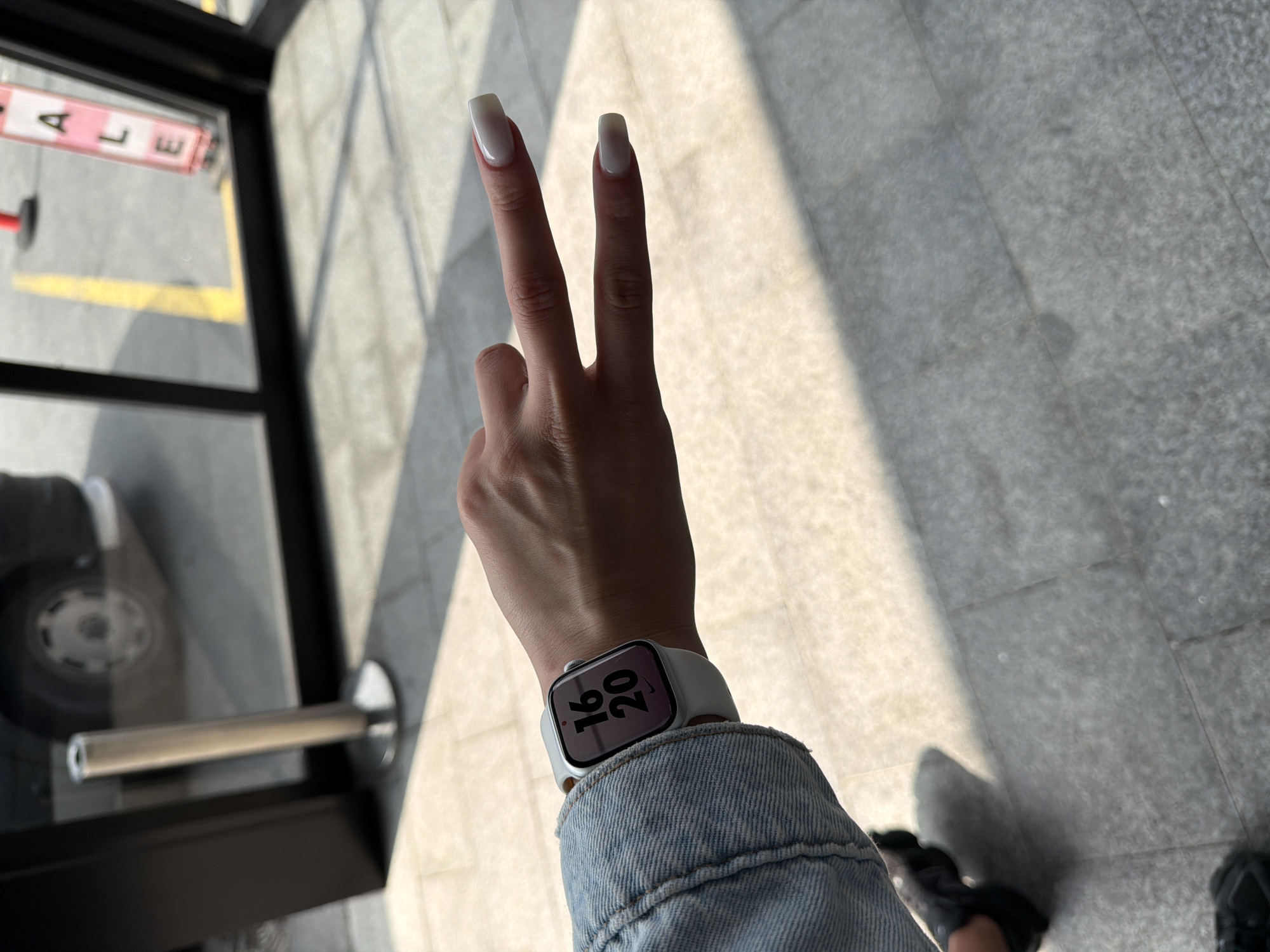 A photograph shows a person's arm and hand, raised to display a peace sign with the index and middle fingers. The fingernails are long and painted with a light color. A smartwatch with a white band is on the wrist, displaying "16:20." The person is wearing a light blue denim jacket. The background consists of gray tiled flooring, a window with sunlight casting shadows, and a glimpse of a vehicle wheel. The letters "ALE" are visible in reverse on a sign seen through the window. The location is Bayrampaşa, Turkey.