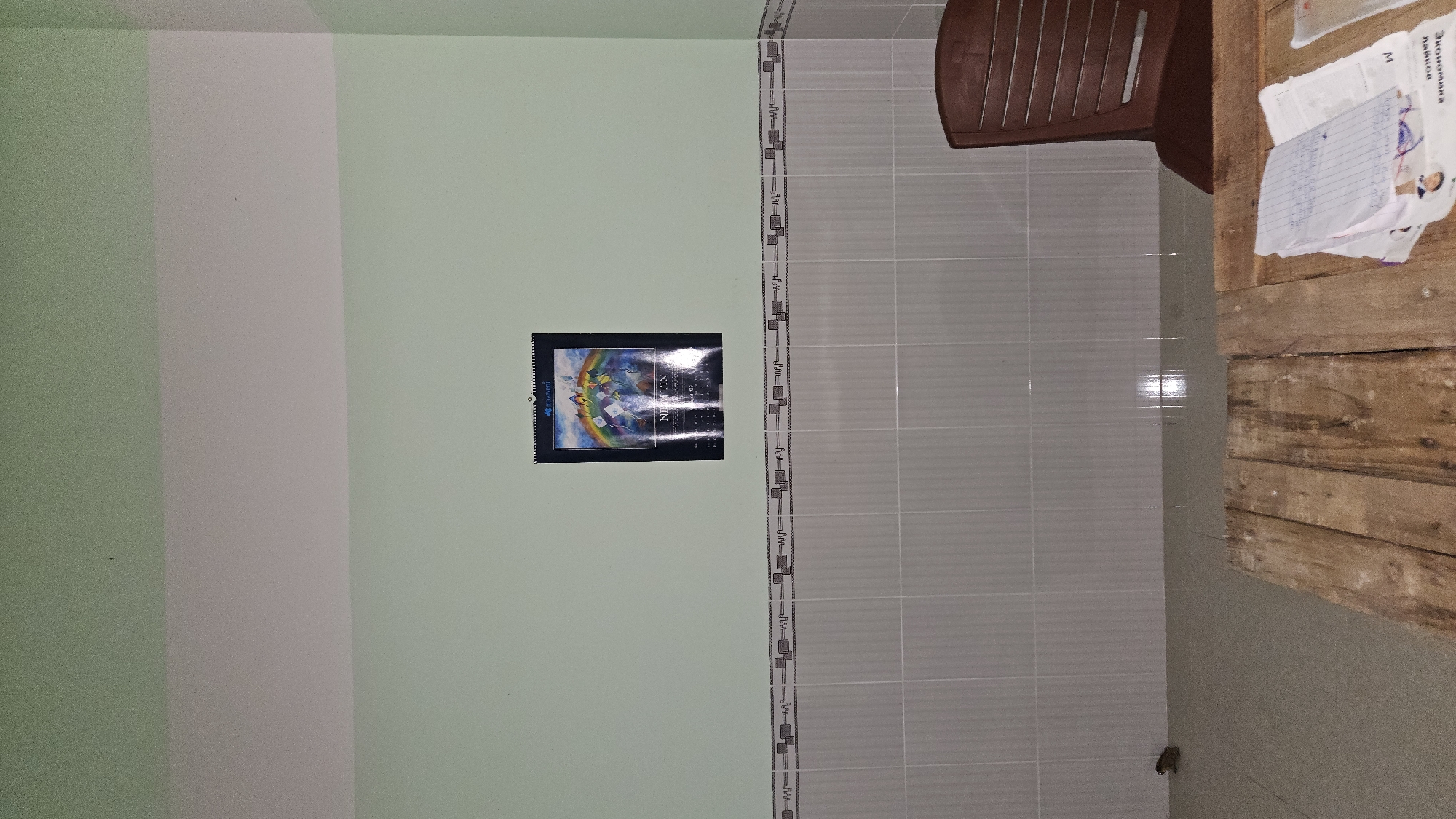 This image, rotated 90 degrees clockwise, captures an indoor domestic space, likely a room within a home.
**Scene/Setting:** The room features contrasting wall treatments. The wall on the left (as viewed in the rotated image) is primarily painted a light, muted green. A vertical, lighter grey or off-white stripe divides this green wall. To the right of this painted section, the wall is covered in light grey rectangular tiles, separated by a decorative patterned border running horizontally across the middle.
**Objects:** A calendar, featuring a vibrant, possibly fantastical illustration (including what appears to be a rainbow or arch), is hung on the light green wall. The word "NEXUS" is clearly visible on the calendar, with "BIORHYTHM" partially seen. In the lower right corner, a rustic wooden table constructed from rough-hewn planks is visible. On this table, several papers are scattered, one containing handwritten notes and another with printed text. The words "Экономика" (Economics) and "данных" (data), written in Cyrillic script, are discernible, along with the letter "M." A brown plastic chair is partially visible, positioned above the table in the upper right. Near the base of the tiled wall, a small, dark, irregular object, possibly a small animal or debris, is present on the floor.
**Activity or Event:** No activity is taking place; the image presents a static view of the room.
**Notable Details:** The lighting in the room appears to be natural or ambient, suggesting daytime. Reflections on the tiled wall indicate a light source. The combination of simple painted walls, tiled sections, and basic wooden furniture gives the space a functional and unpretentious character. The presence of text in Cyrillillic on the papers is a notable detail for a setting in Cam Đức, Vietnam, potentially indicating educational materials or a cultural connection.