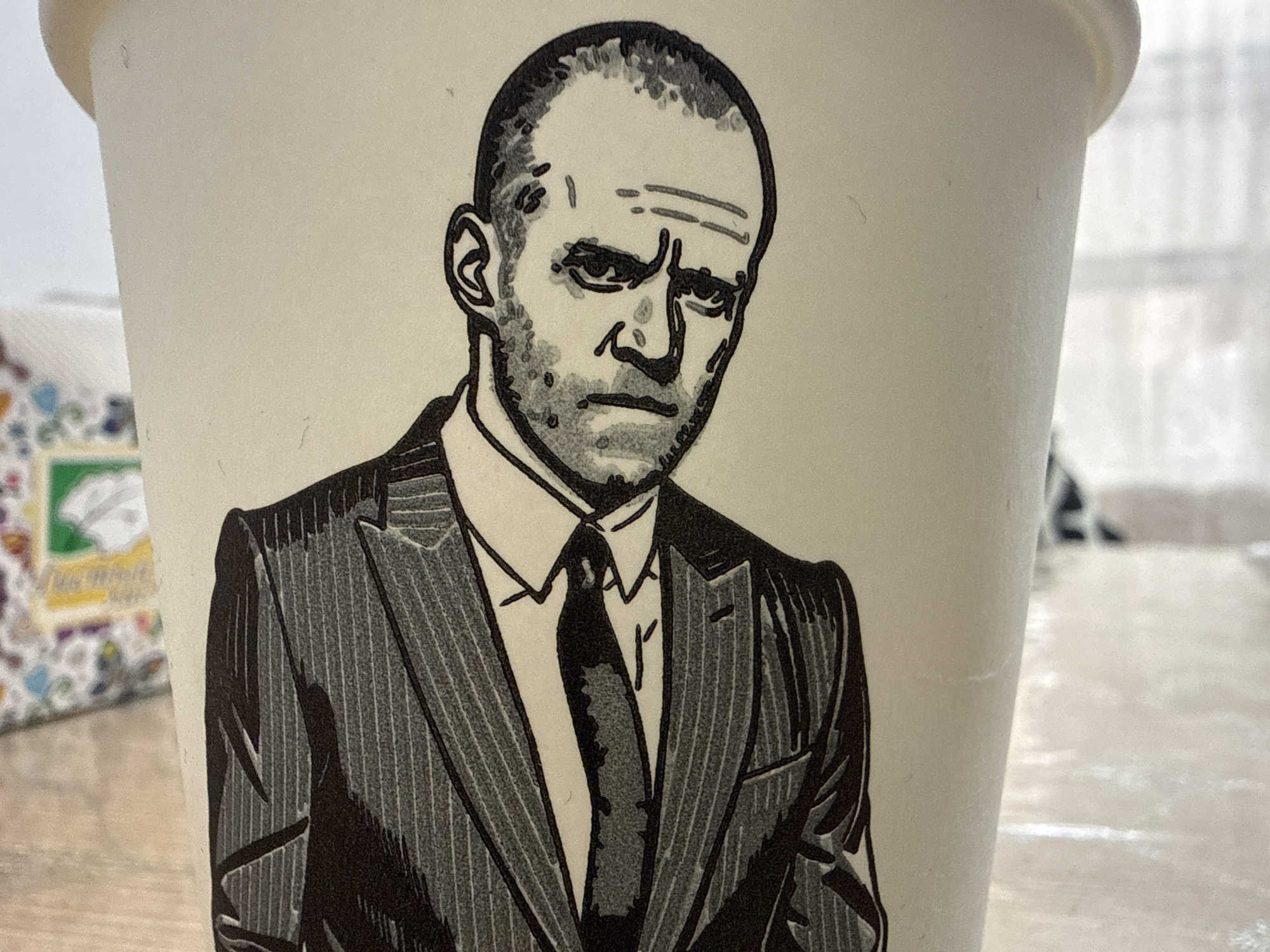 A close-up, eye-level, medium shot captures a black and white sketch of a stern-faced bald man on a paper cup. The man wears a suit and tie, with heavy shading and lines depicting his furrowed brow, intense eyes, and stubble. The cup is positioned on a light-colored surface, likely a table, with a blurred background that suggests an indoor setting. A portion of another patterned object is visible to the left of the cup. The lighting is neutral and even. There is no indication of the city of Beslan, Russia, or any specific geographical location.