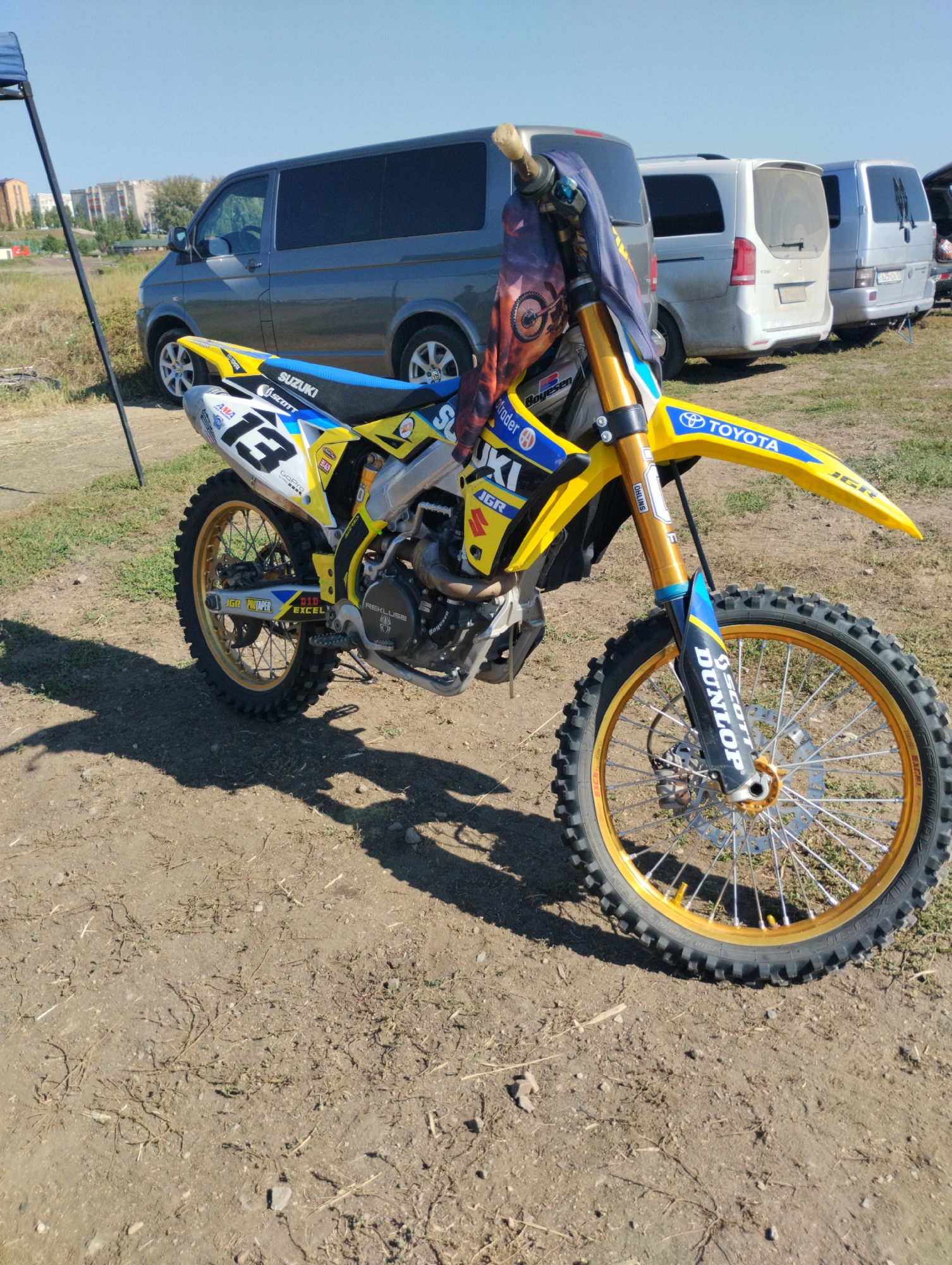 A yellow and blue Suzuki dirt bike with the number 13 prominently displayed on its side is parked on a dirt surface, likely a dirt track or parking area, on a clear day. The bike has gold rims on both wheels and a black tire with aggressive treads. The handlebars are adorned with a patterned cloth featuring a motorcycle wheel design. In the background, several vans, including a dark grey Volkswagen van and two lighter-colored vans, are parked. The environment appears to be outdoors, with dry grass and some sparse vegetation visible. In the far distance, buildings and trees suggest an urban or semi-urban setting. The scene is consistent with an outdoor recreational area for dirt biking, potentially located in or near Kostanay, Kazakhstan, given the typical appearance of urban landscapes and vehicle types found in the region.