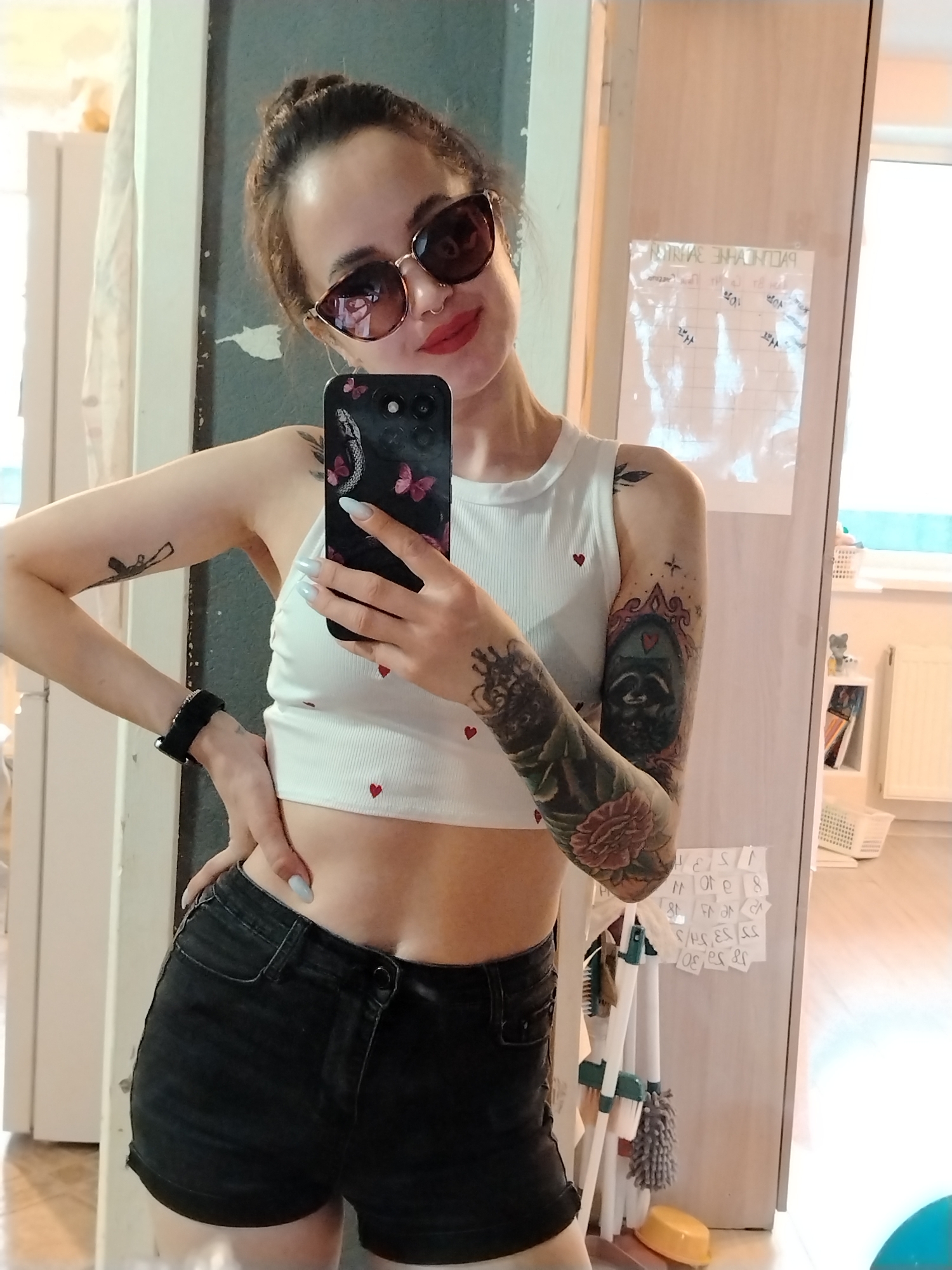 A woman is standing in front of a mirror, taking a selfie. She is wearing sunglasses, a white crop top with red hearts, and black denim shorts. She has several tattoos on her arms. She has her hair up in a bun. Her phone has a black case with a snake and butterflies. The background includes a door, a calendar, cleaning supplies, a bookshelf, and a window. The location is not specified as the city of Noviy, Russia.