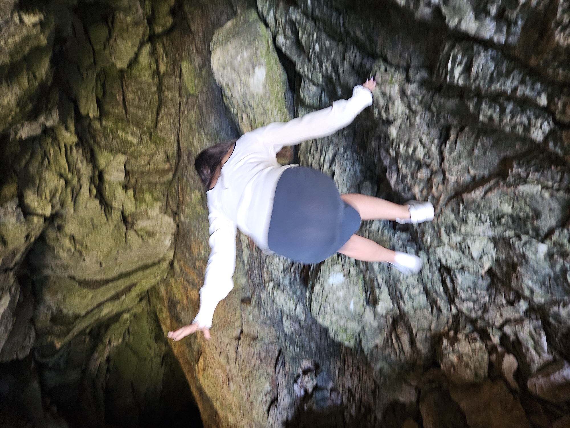 A blurry, skewed, low-angle shot depicts a person falling or leaping in a dark, rocky environment, possibly a cave. The person, viewed from the back, wears a white long-sleeved shirt, a dark skirt or shorts, and white shoes. Arms are outstretched. The surrounding environment consists of dark, textured rocks. The location is unknown.