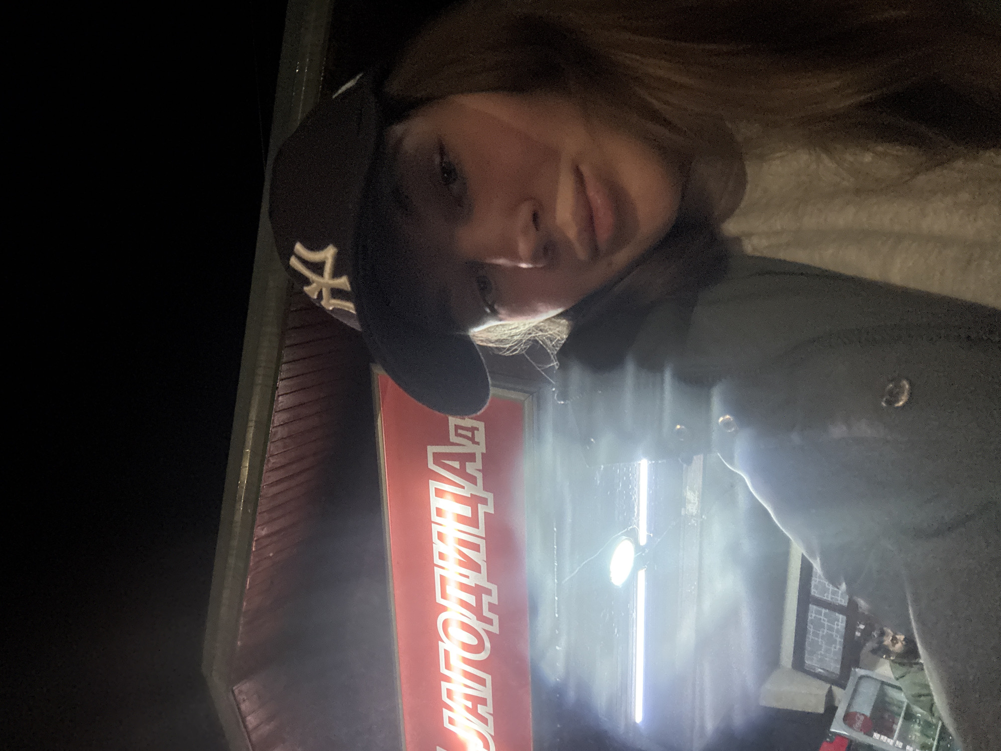 A woman with long brown hair is posing for a selfie in front of what appears to be a commercial building. She is wearing a black baseball cap with a "NY" logo. The background is dark, likely taken at night. Part of a red sign is visible, displaying Russian text "УГОДИЦА". The photograph includes a portion of the building's facade, suggesting a street-level view. The woman is positioned slightly to the right in the frame. The bright lights on the store front are visible.