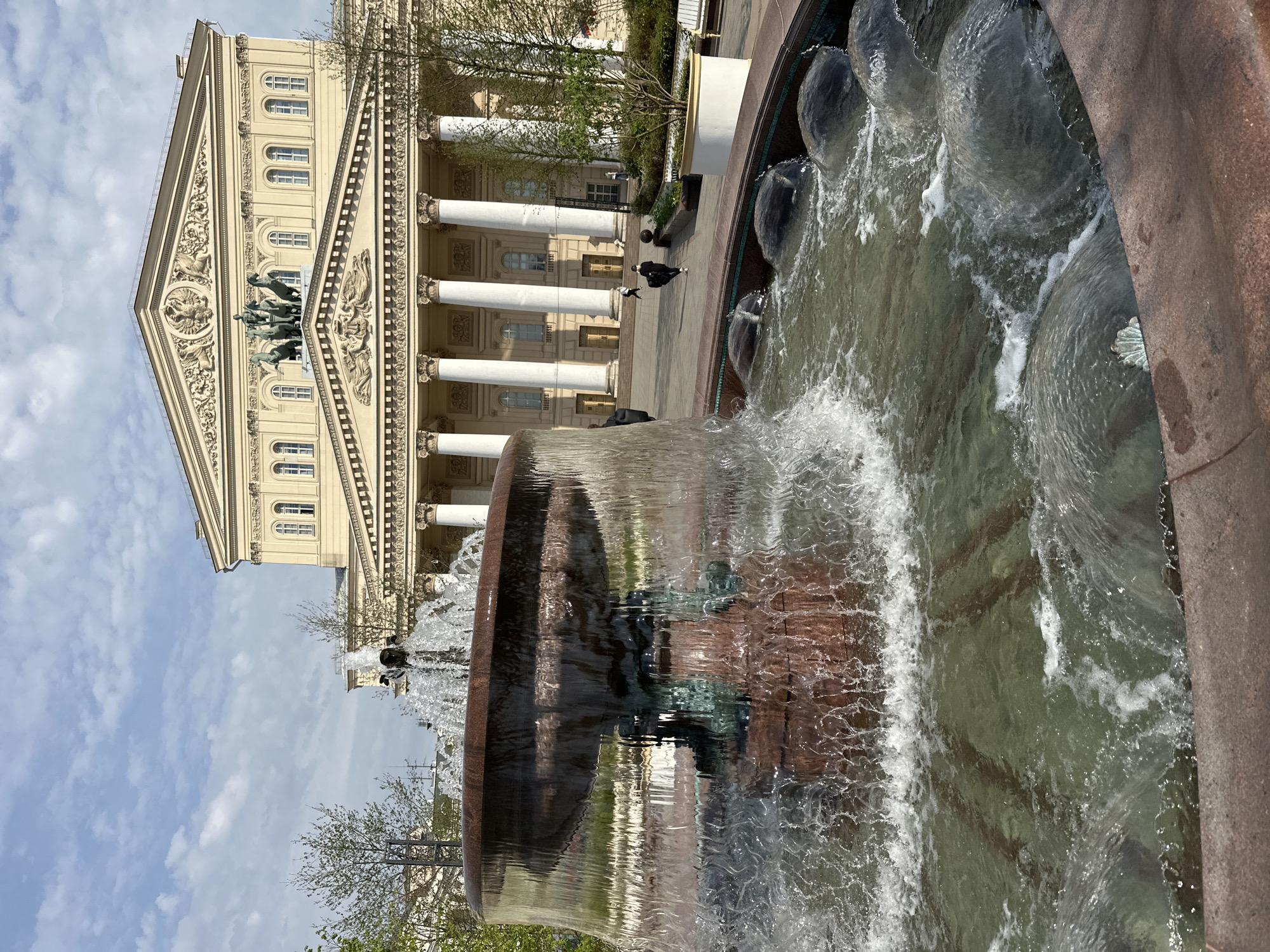 The image shows a fountain in front of the Bolshoi Theatre in Moscow, Russia. The fountain is multi-tiered with water cascading down. The Bolshoi Theatre is in the background, showcasing its neoclassical façade with columns and a sculpture of Apollo. The sky is partly cloudy. People are seen walking in front of the theatre.