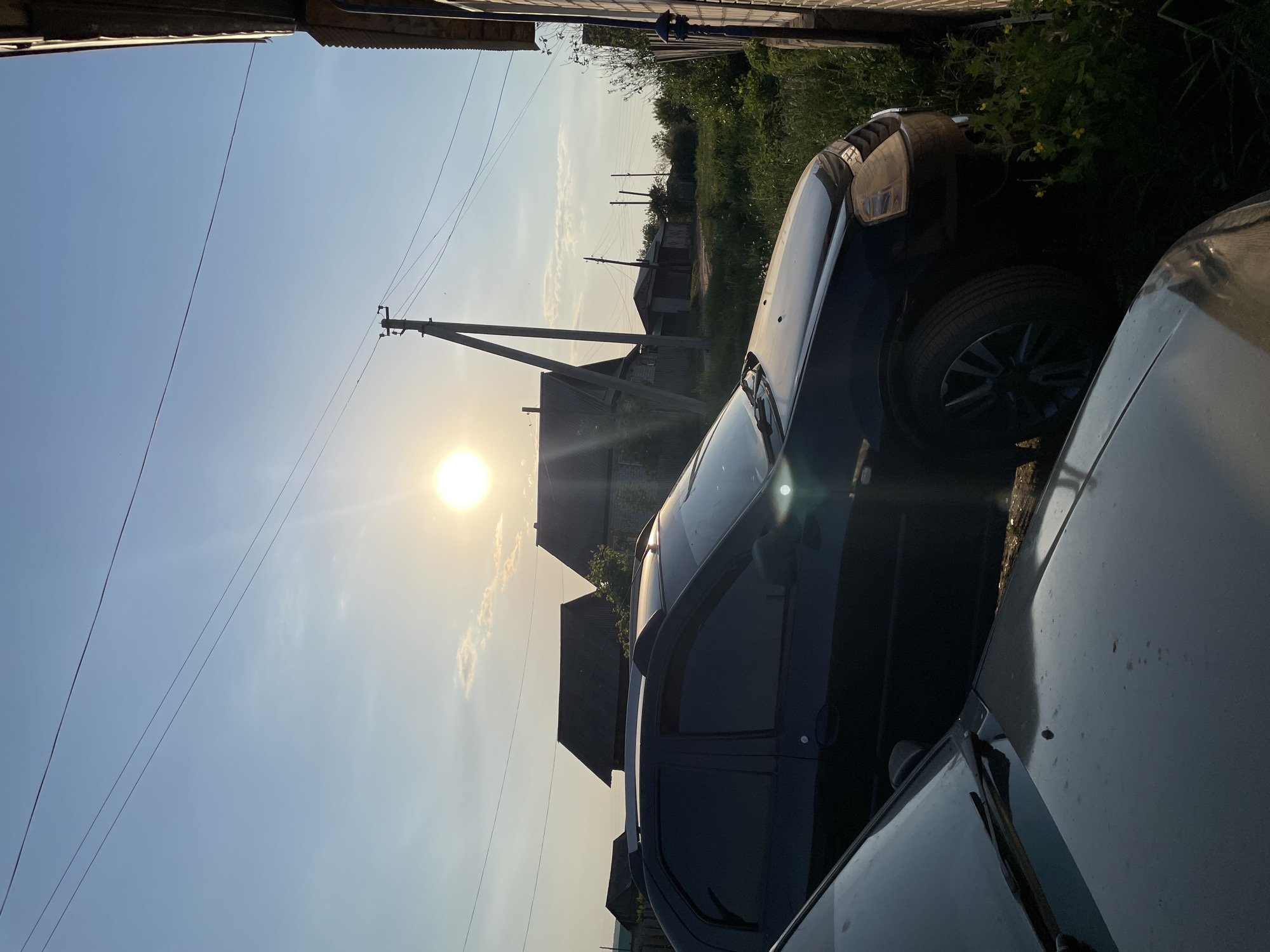 The image shows a street scene in Кыйлуд, Russia, with two parked cars in the foreground. A dark-colored car is parked next to a light-colored one. In the background, there are houses, power lines, and vegetation. The sun is visible in the sky.