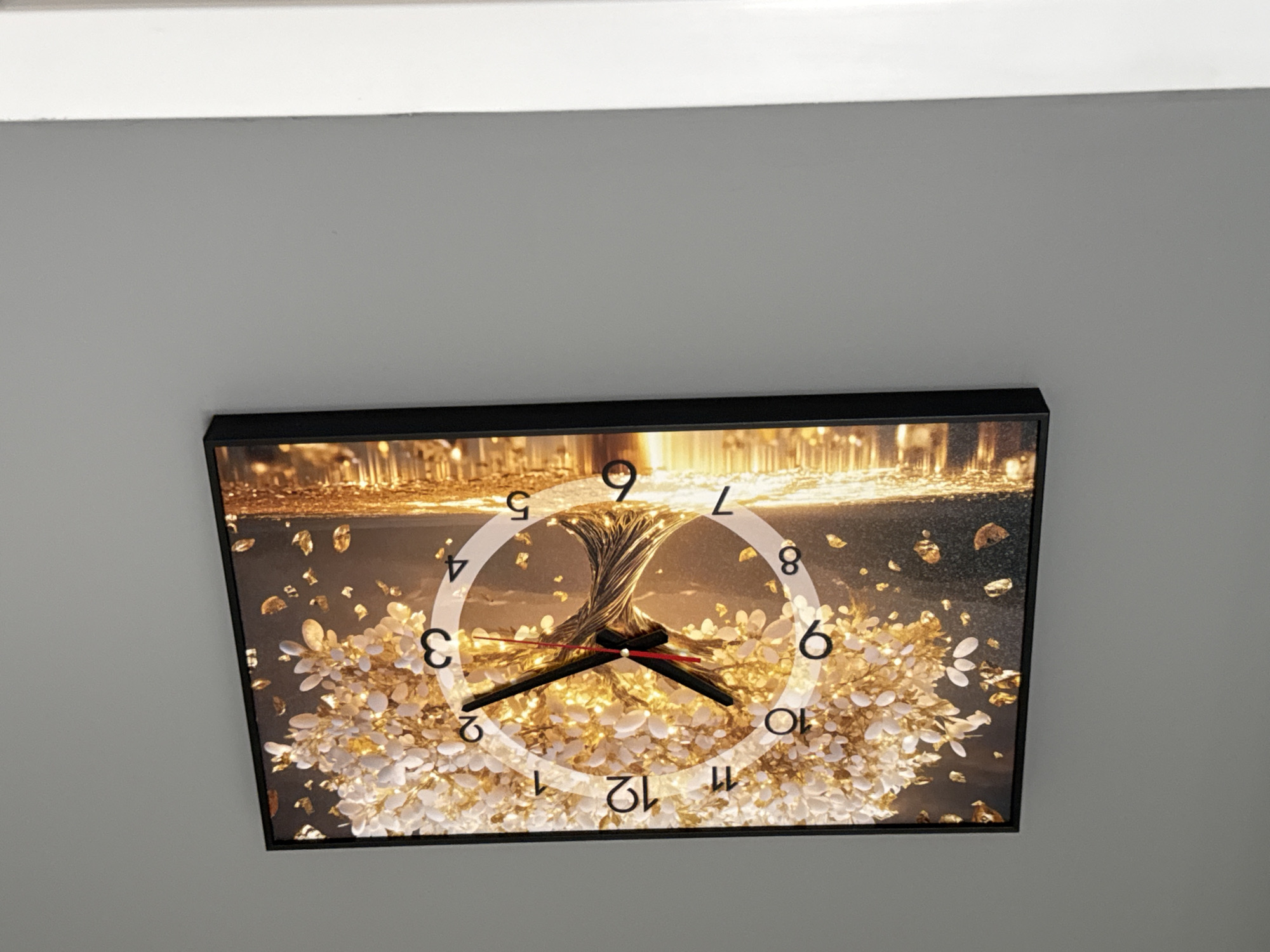 A rectangular wall clock with a black frame hangs on a plain gray wall. The clock face features a detailed image of a golden tree with luminous leaves and a swirling trunk, set against a dark, reflective background. The numbers 1 through 12 are visible around the circumference of the clock face, with a white circle as a guide. Black hour and minute hands, along with a thin red second hand, are positioned on the clock face. The hour hand points between the 1 and 2, the minute hand points to the 2, and the second hand points to approximately the 10. The clock appears to be digital, with the image superimposed onto its face. No people or specific Yekaterinburg landmarks are visible in the image.