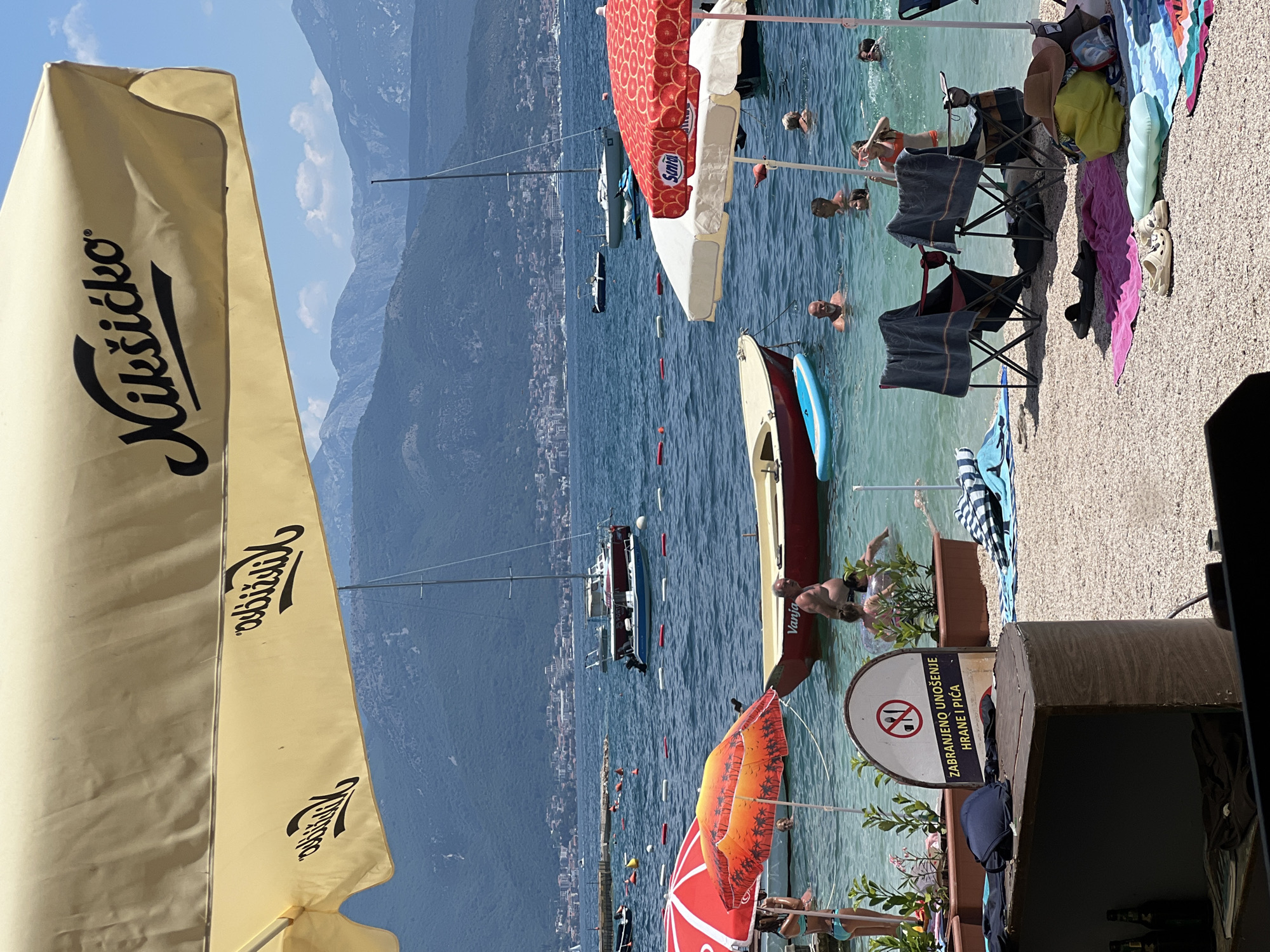 The image depicts a sunny day at a beach in Baošići, Montenegro. In the foreground, a large, yellow umbrella with the "Nikšićko" logo provides shade. The water is clear and turquoise, with several boats visible in the background, including sailboats and smaller motorboats. People are enjoying the water, swimming and relaxing. Beach chairs and towels are scattered on the pebbled shore. A sign with a prohibition symbol indicating no food or drinks is also visible. The backdrop features lush green hills and mountains, characteristic of the Bay of Kotor.