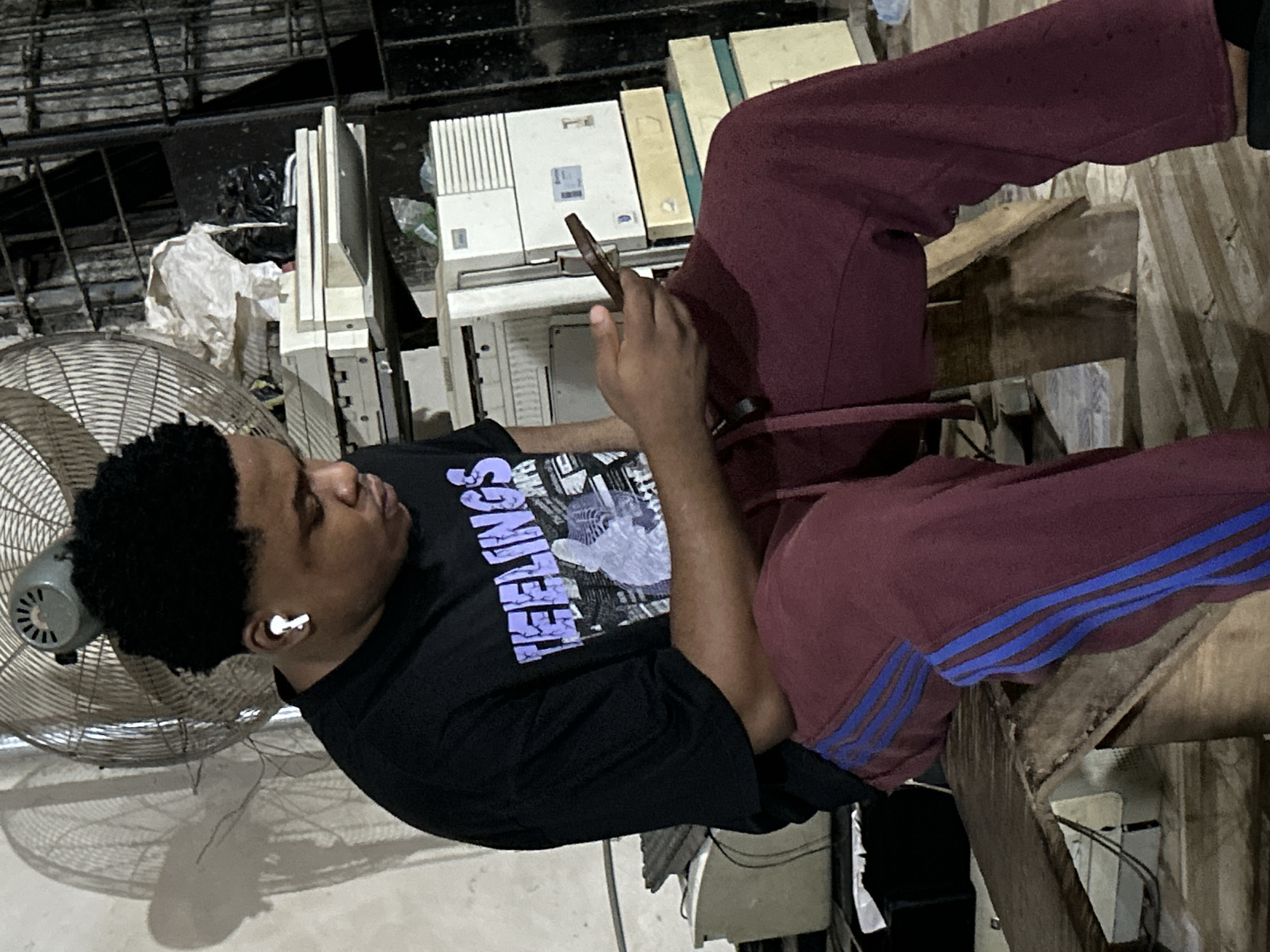 A young Black man is seated on a wooden chair in what appears to be an indoor, cluttered space. He is wearing a black t-shirt with a graphic design and the word "TEENAGE" printed on it, as well as maroon-colored Adidas track pants with blue stripes down the sides. He has white wireless earbuds in his ears and is looking at a phone he holds in his hand. To the left, a large, old-fashioned metal fan is visible. Behind him, there's a stack of old computer monitors and other electronic equipment. The environment appears to be a workshop or storage area in Ihiagwa, Nigeria.