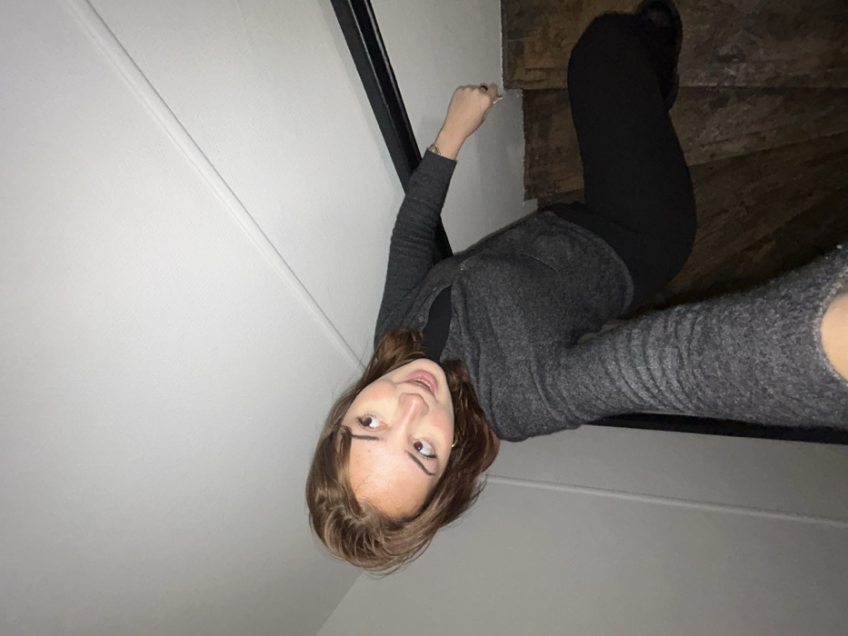 A single female individual with medium brown hair is depicted in an indoor setting, seated on a dark wooden staircase. She wears a dark gray knit cardigan over a black top and black trousers. Her head is tilted to her right, and her gaze is directed upward and slightly to the right with a subtle smile, indicating direct interaction with the camera held in her extended left hand. A ring is visible on her right ring finger, and a thin bracelet on her right wrist. Her right hand rests on the step above her. The surrounding environment consists of white walls with a dark handrail and a white ceiling featuring molding. The image is captured from a low-angle perspective. The specific location within Strasbourg, France, cannot be determined from the visual information provided.