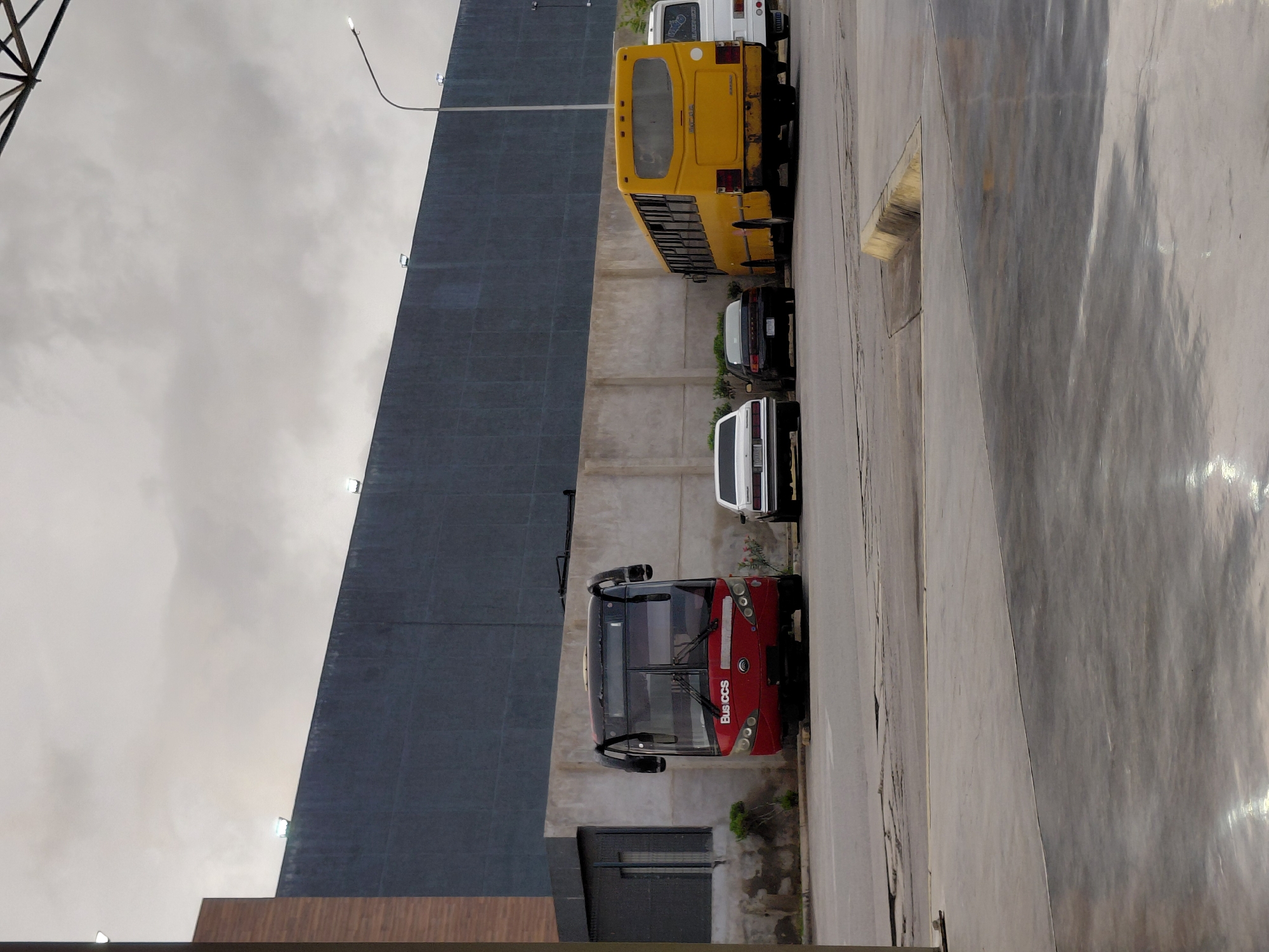 In Juan Griego, Venezuela, under a cloudy sky, a line of parked vehicles is visible. In the foreground, a light gray concrete surface shows reflective puddles. Behind this, a paved area is cracked in places. Further back, a red bus with "BusCCS" written on its front is parked. To its right, a white sedan is parked, followed by a black sedan. The largest vehicle in the image is a yellow bus, positioned behind the sedans, with its rear facing the viewer. To the right of the yellow bus, a white van is parked. A tall, dark gray wall forms the backdrop for the parked vehicles. A street light stands to the right of the yellow bus. A section of a brown and gray building is visible on the far left.