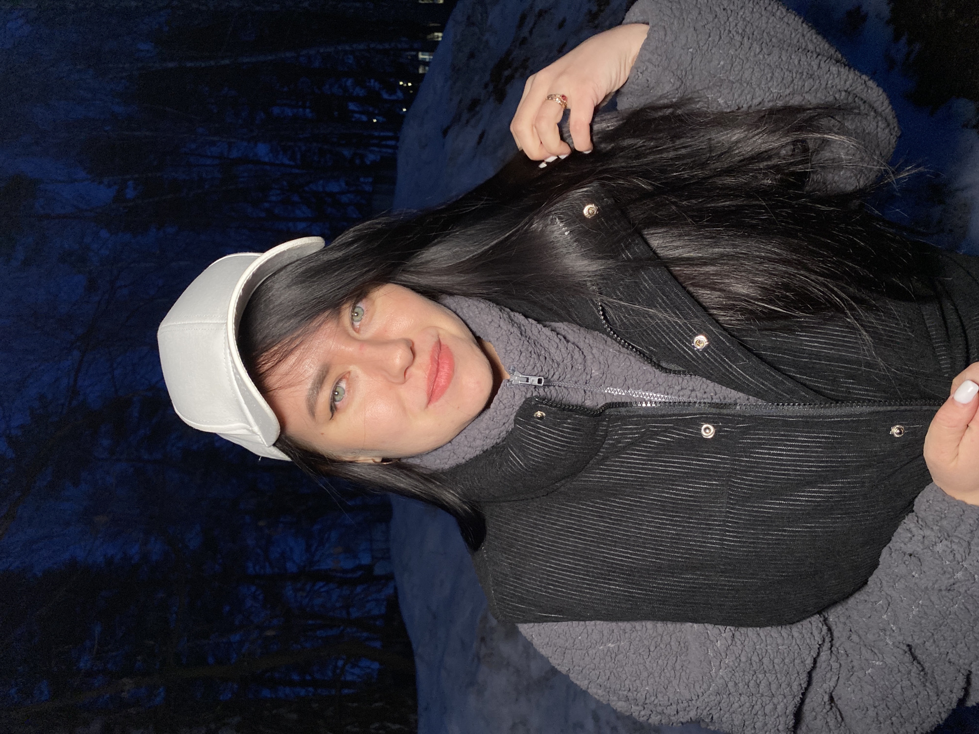A woman with long black hair and fair skin is wearing a white baseball cap and a dark-colored vest over a grey fleece jacket. She is smiling slightly and looking at the camera. She has a ring on her left hand. The background shows trees against a dark blue sky, possibly indicating nighttime or twilight. The location is possibly Chaikovsky, Russia, based on the prompt. The ground has patches of snow.