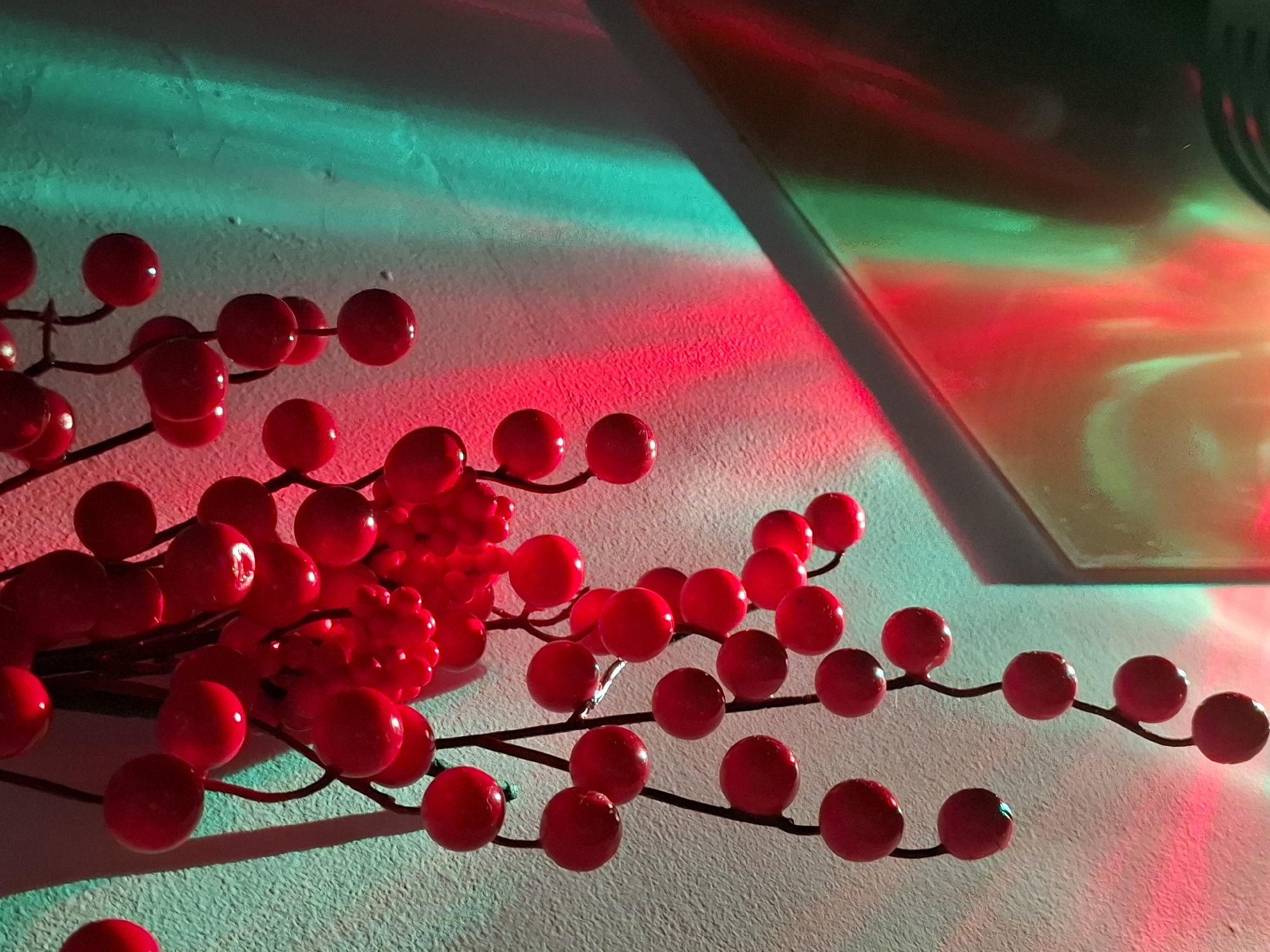 The image shows a bunch of artificial red berries hanging from a dark stem against a textured, off-white wall. The wall is illuminated by red and green lights, casting distinct colored shadows and reflections. The lighting creates a festive or decorative atmosphere. There are no people visible in the image. The scene appears to be an indoor decoration, possibly for a holiday. The specific location in Zemun Urban Municipality, Serbia, cannot be determined from the visual information provided in the image.