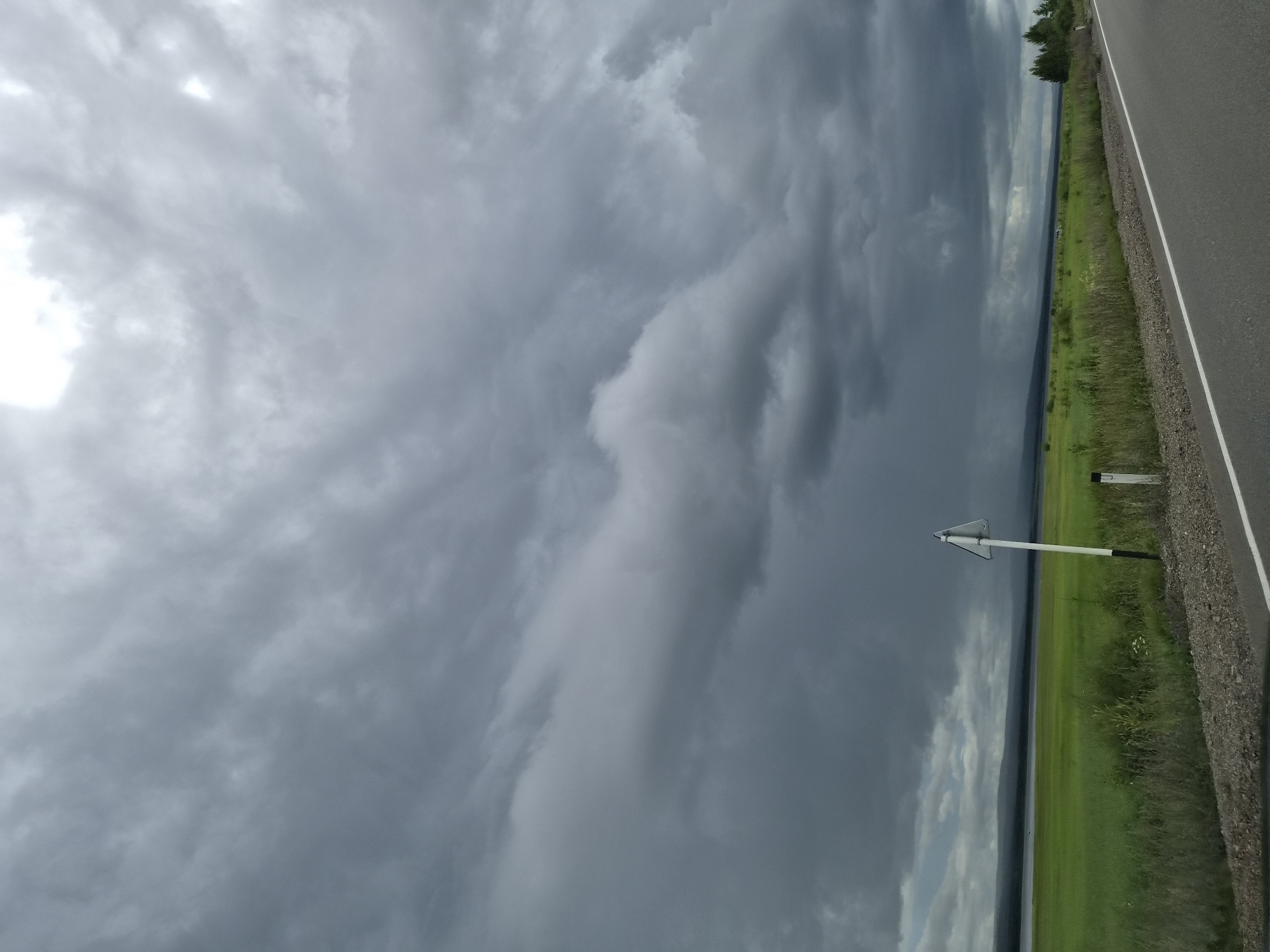 The image shows a view from the side of a road in Ivan-Ozero, Russia. The road is paved and has a white line marking the edge. To the left of the road is a field of green grass extending towards a body of water in the distance. A dark, cloudy sky with dramatic cloud formations hangs overhead. There is a white triangular road sign on a pole near the side of the road. The overall scene depicts a somewhat overcast and potentially stormy day in a rural or semi-rural landscape.