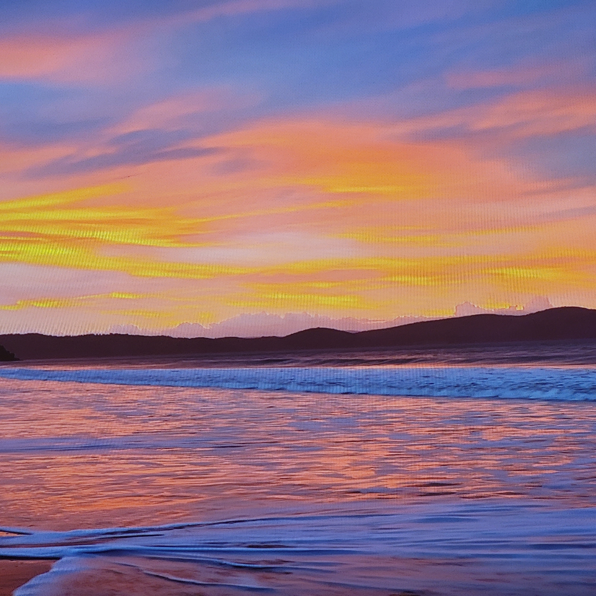 The image depicts a beach scene at sunset. The sky is a mix of blue, orange, and yellow hues, with clouds stretched horizontally. In the midground, there is a dark silhouette of hills. The ocean reflects the colors of the sky, with waves near the shore. The foreground shows the wet sand and advancing tide with faint reflections of orange-pink colors. There is no evidence in the image to suggest that this scene is in Yerevan, Armenia, or any other city. The location cannot be determined.