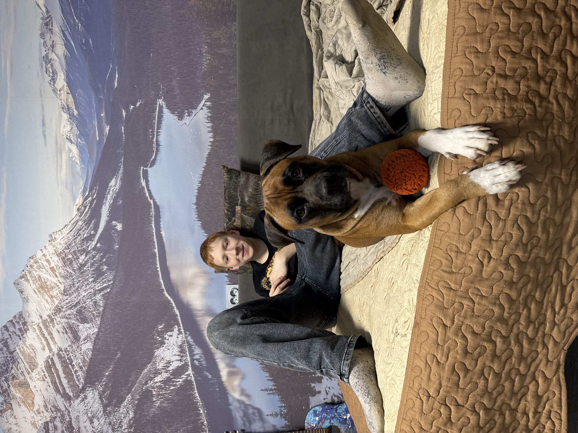 An indoor portrait shows a young boy and a dog relaxing together, likely on a bed or sofa. The setting features a large wall mural depicting a snowy mountain range with a lake and dense pine forests, creating a natural backdrop.
The boy, with short reddish-brown hair, is smiling faintly at the camera. He wears a dark t-shirt with a colorful graphic and dark grey jeans, paired with white socks speckled with blue or grey. He is seated with his legs bent.
Beside him, a fawn-colored Boxer-type dog with a black muzzle and white markings on its chest and paws lies facing the camera. An orange, textured ball rests near its front paws.
Underneath the subjects, a quilted blanket or bedspread in cream and brown tones is visible, with the brown section featuring a distinct wavy pattern. On the wall behind the boy, a white double electrical outlet can be seen. A small part of a blue and white patterned guitar is visible on the floor near the boy's feet. The overall atmosphere is relaxed and comfortable, suggesting a casual moment shared between the boy and his pet in a home located in Chaikovsky, Russia. No visible text is present.