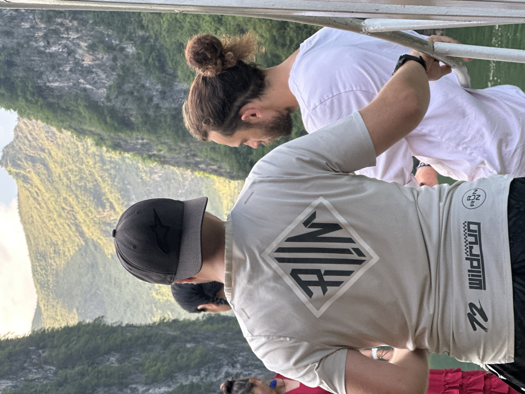 Three people stand on a vessel near Đồng Văn Commune, Vietnam. A person with hair in a bun leans over the railing, wearing a white t-shirt. Another person, facing away from the camera, wears a baseball cap and a tan t-shirt with a diamond logo and the word "Wildrun" on it. A third person is visible between them, and a fourth person is visible in the bottom of the frame. Lush, green mountains rise in the background. The image appears to have been rotated.