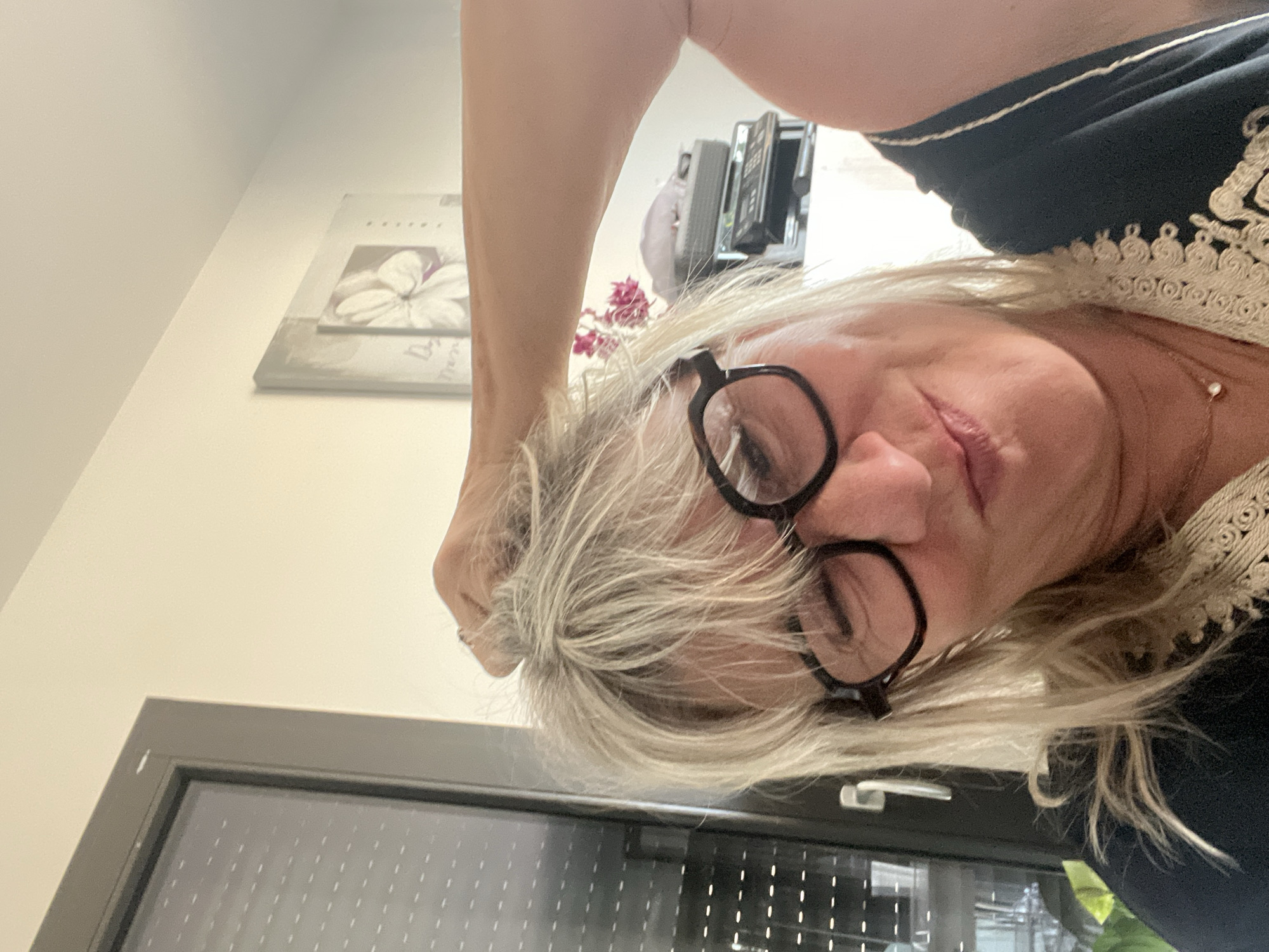 A close-up, overhead shot shows a woman with blonde hair and dark-rimmed glasses looking at the camera. She has her right arm raised, and her hand is in her hair. The woman is wearing a dark top with a decorative trim. The background shows a window with blinds, a wall with a framed picture of a flower, and a piece of electronic equipment on a shelf. The scene appears to be indoors, possibly in a residential setting. The image quality is slightly blurry, suggesting it may have been taken with a phone camera.
