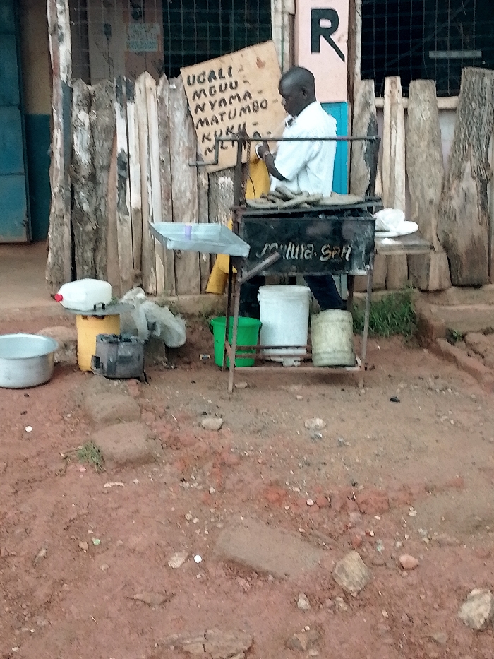 The image shows a man cooking in front of a small stand in Kivaywa, Kenya. The stand is made of a wooden fence with a metal frame and a grill. The man is wearing a white shirt and is standing next to the grill. There is a sign behind him that says "UGALI, MGUU, NYAMA, MATUMBO, KUKU, ML" in a yellow color. The man appears to be preparing food for customers. The scene is located at latitude 0.562 and longitude 34.805 in the city of Kivaywa, Kenya. There are a number of containers, some of which are full of water. The area is mostly dirt, with a few rocks and some grass.