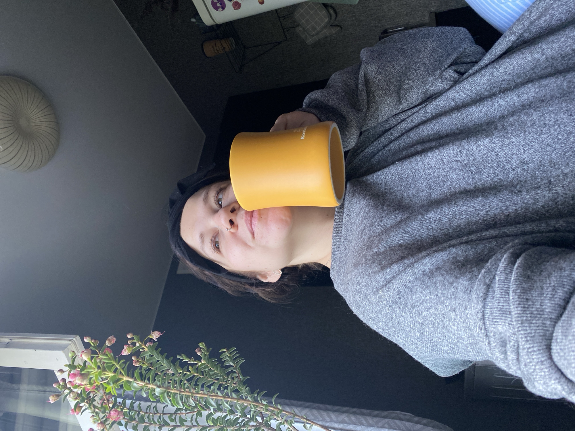 A woman is holding a yellow mug up to her face in an indoor setting. The woman is wearing a grey sweatshirt and a black headband. In the background, there is a window with a plant in front of it, a ceiling light, and a glimpse of other objects on a surface. It is impossible to determine the location as Chaikovsky, Russia based on the image.