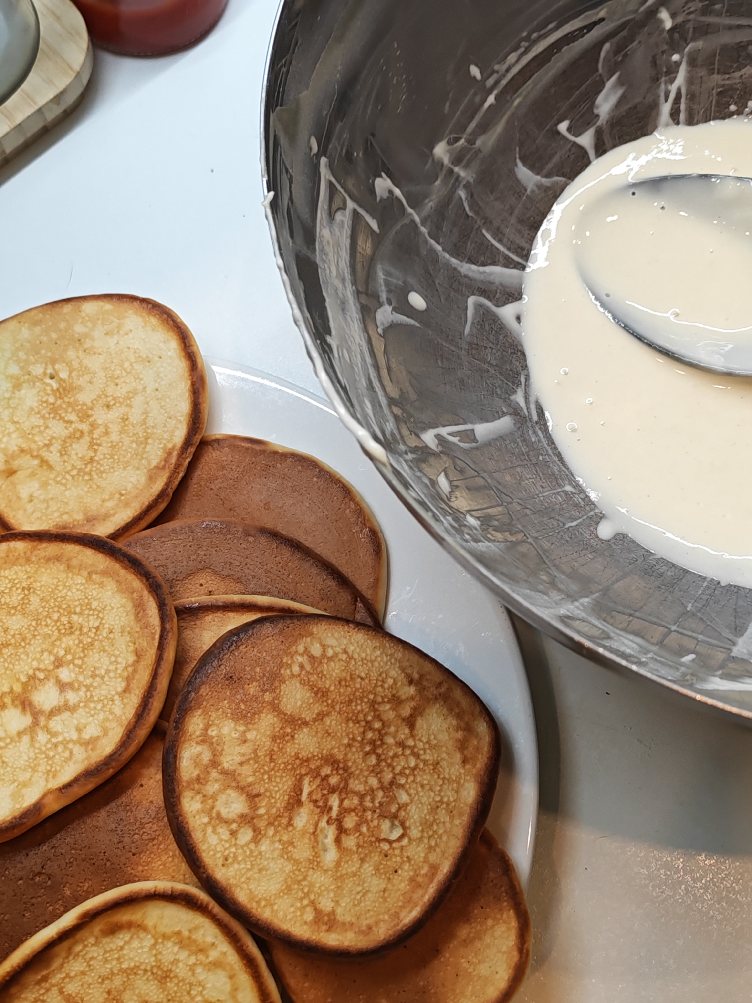 A plate of golden brown pancakes is shown in close-up, next to a metal bowl containing pancake batter and a spoon. The pancakes are stacked on a white plate and appear to be small and round. The batter in the bowl is thick and creamy, with some white streaks from where the batter has been scraped from the sides. The scene is set on a white countertop, and in the background, a wooden object and a red bottle are partially visible. The image is well-lit, with the natural light casting shadows on the objects. The setting is likely a kitchen, and the food preparation is in progress. There is no indication of the location being in Bataysk, Russia.