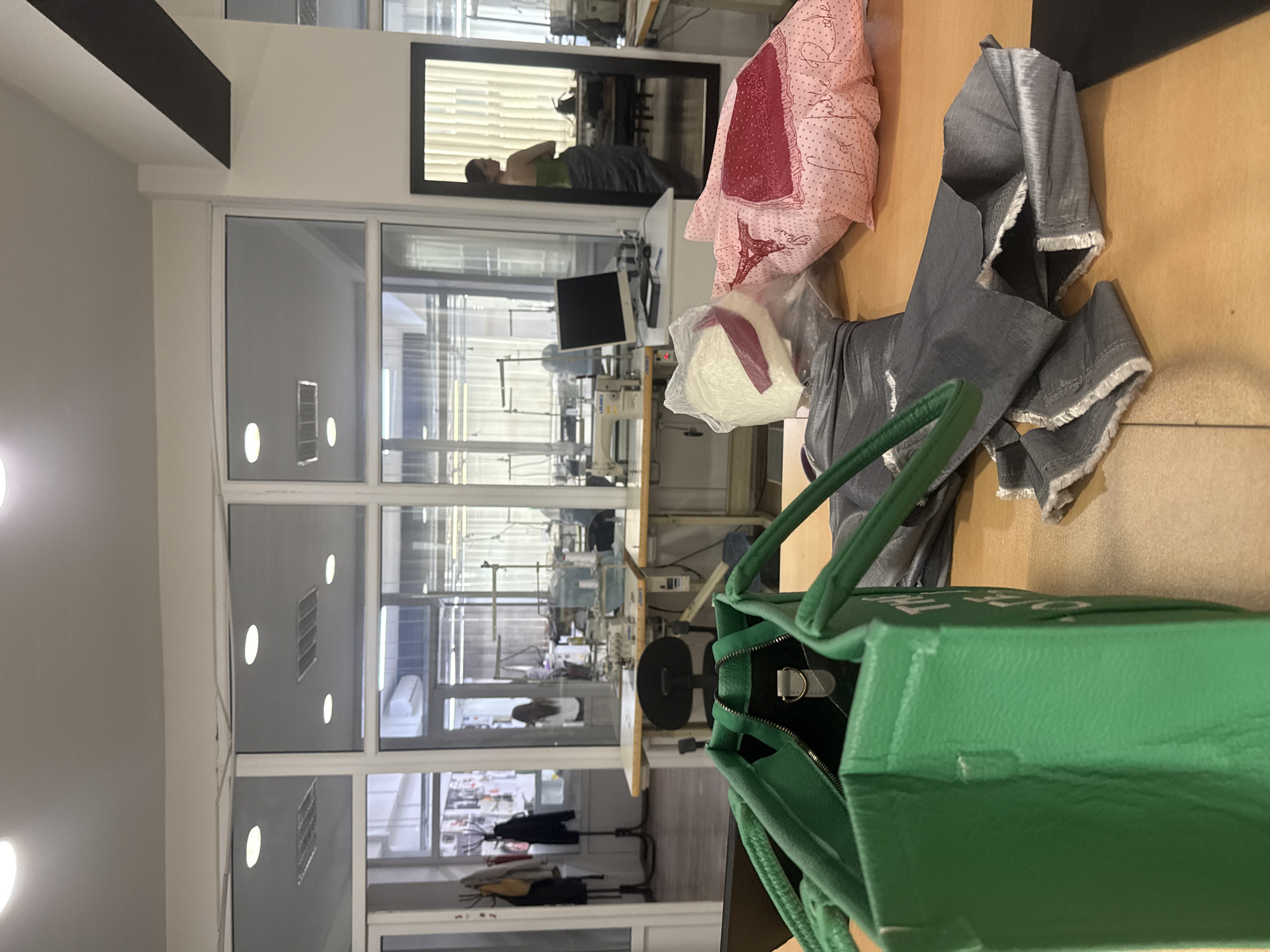 A view of the interior of a textile workshop, possibly in Yerevan, Armenia. The workspace features large windows with multiple panes, allowing ample natural light. In the foreground, a green tote bag sits partially on a wooden table, obscuring some of the objects behind it. A pink patterned fabric, a bundle of white material, and a piece of gray textile are positioned on the table next to the bag. A mirror hangs on the wall, reflecting a person in a green shirt, partially obscured by the items on the table. In the background, multiple sewing machines can be seen on tables, indicating a manufacturing or tailoring environment. The workshop appears to be in active use, with various tools and equipment in place.