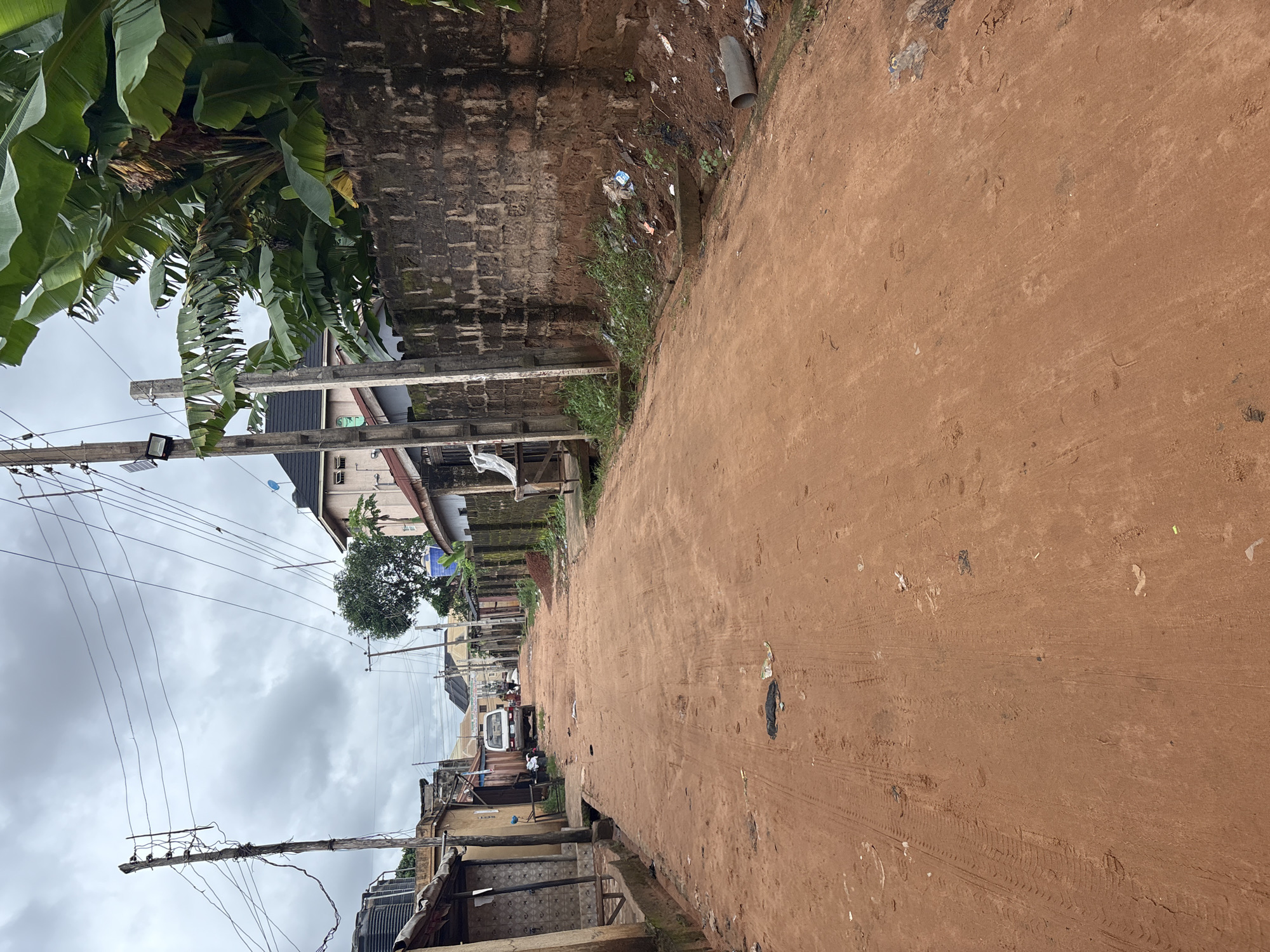 The image depicts a street scene in Nkpor, Nigeria. The street is unpaved, with a red dirt surface showing tire tracks. On the left side of the street, there are single-story buildings with corrugated metal roofs and some structures made of cinder blocks. Overhead, electrical wires are strung between wooden poles. Further down the street, a white van is parked. On the right side of the street, a tall, rough brick wall runs along the property line, topped with banana trees and other foliage. Debris, including plastic waste and a detached concrete pipe, is visible along the base of the wall and on the edge of the street. The sky is overcast with gray clouds.