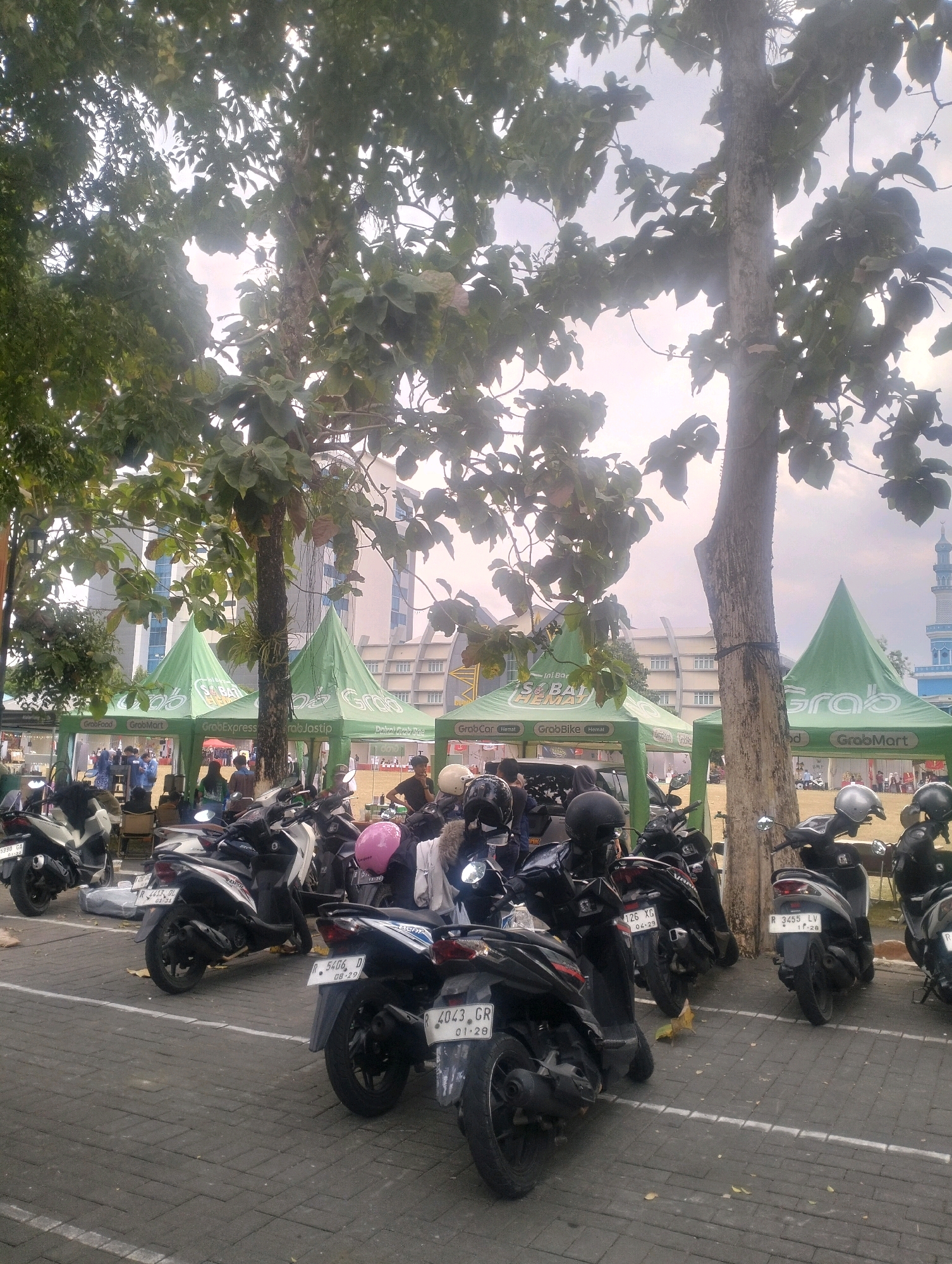 The image depicts an outdoor scene in Karangsoka, Indonesia, characterized by a parking lot filled with parked motorcycles. Several green tents, branded with "Grab" logos, are set up, suggesting a promotional event or service point. The tents are spaced along a paved area, with trees providing partial shade. In the background, buildings with modern architecture are visible, including what appears to be a mosque with a blue dome and minaret. People are present, some standing near the tents and others possibly involved in the activities within or around them. The motorcycles are mostly scooters, parked in rows. The overall environment suggests a public gathering or commercial activity taking place outdoors.