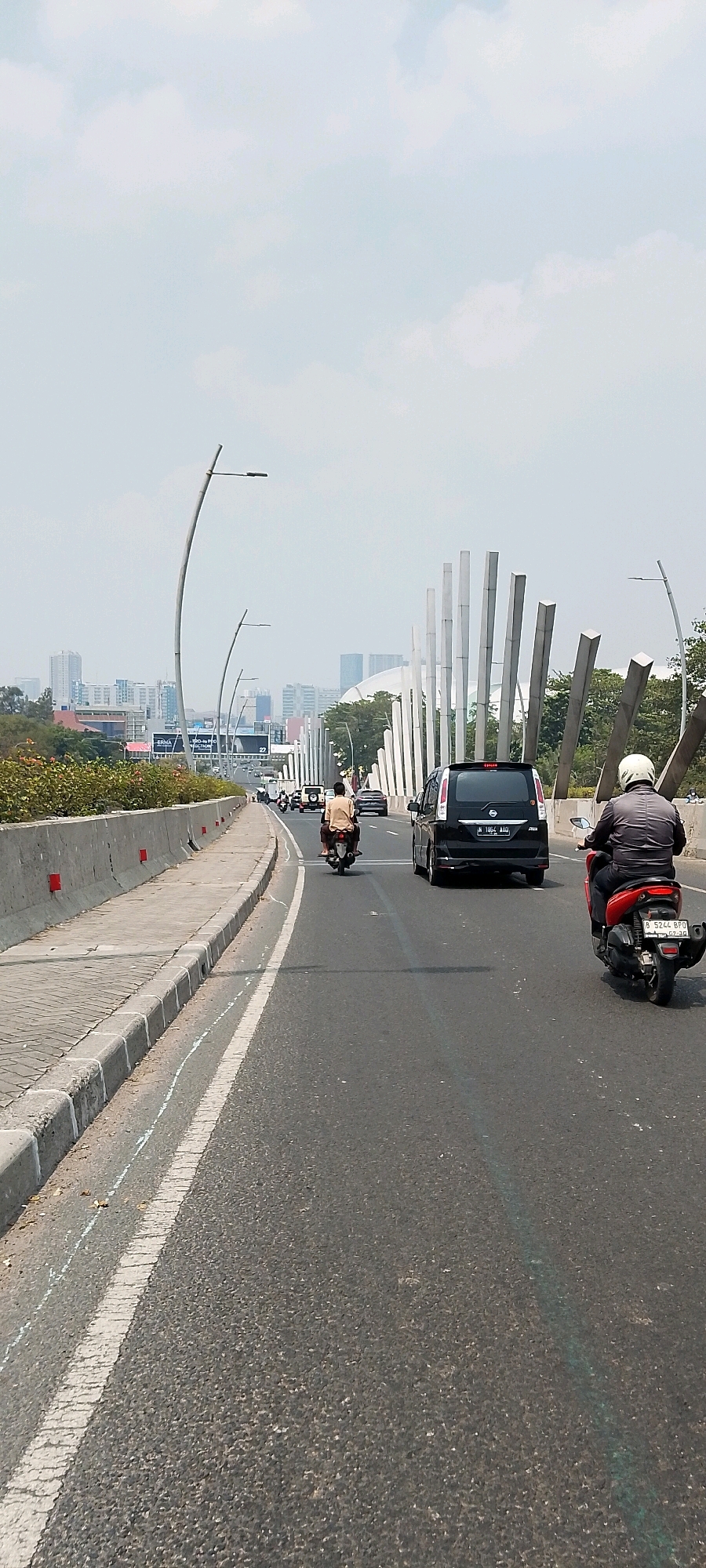 This image captures a street scene in Bekasi, Indonesia, likely on an elevated road or highway. In the foreground, a dark-colored car with license plate B 1894 AAO is driving on the right side of the road. To its right, a person on a red motorcycle, wearing a grey jacket and white helmet, is also traveling in the same direction. Further ahead, another motorcycle with two occupants, one in a light-colored shirt and the other in the back, is visible in the lane. The road is paved and marked with white lines. On the left side of the road, there is a sidewalk with a concrete barrier and decorative bushes. In the background, there is a cityscape with various buildings, including skyscrapers, and an architectural structure with tall, slender white pillars. The sky is overcast with a light haze. The overall impression is of a busy urban environment with moderate traffic.