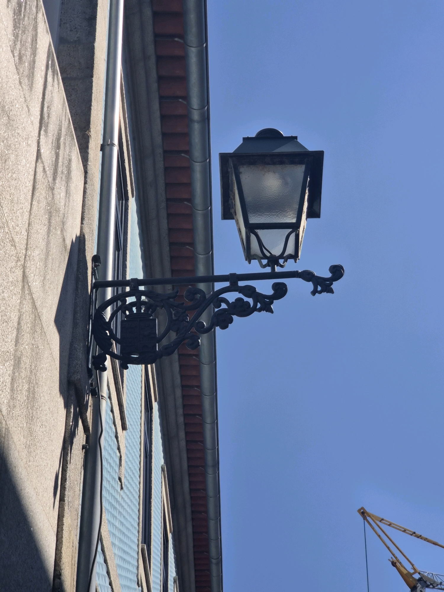 This is a low-angle, close-up, outdoor, daytime photograph of a vintage wrought-iron street lamp attached to the side of a building. The building has a textured, light-colored stone facade on the left and a section with pale blue, patterned siding to the right of a vertical downspout. Above the siding, there is a row of red tiles and a gutter. The street lamp itself is dark, ornate, and extends horizontally from the wall. It features a square, multi-paneled glass lantern with a dark top. Decorative scrollwork is visible on the bracket supporting the lantern, including a circular element that appears to be a coat of arms. The background is a clear, bright blue sky, with a yellow construction crane visible in the far lower right corner. The image was taken in Barcelos, Portugal.