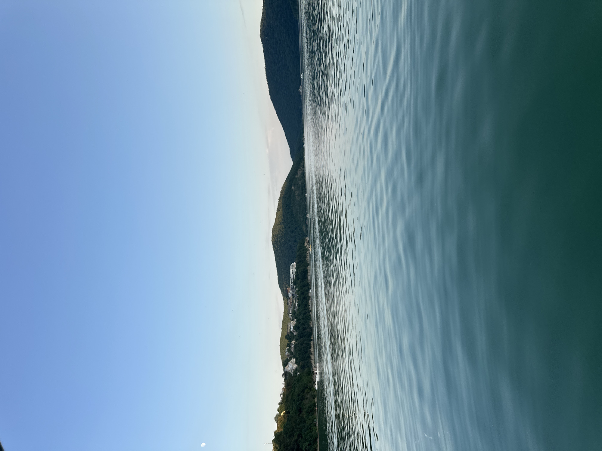 The image depicts a scenic view of a body of water, likely a lake or sea, with a lush, tree-covered hillside bordering it. The water's surface is rippled, reflecting the light and creating a dynamic texture. The hillside is dotted with scattered white buildings, suggesting a settlement or resort area. The sky is a clear, pale blue, transitioning to a lighter hue near the horizon. The overall impression is one of tranquility and natural beauty. A small, crescent moon is visible in the sky. The image appears to have been taken during daylight hours, possibly in the late afternoon or early evening, given the soft lighting. There is no visible text in the image. The location is identified as Abrau-Dyurso, Russia.