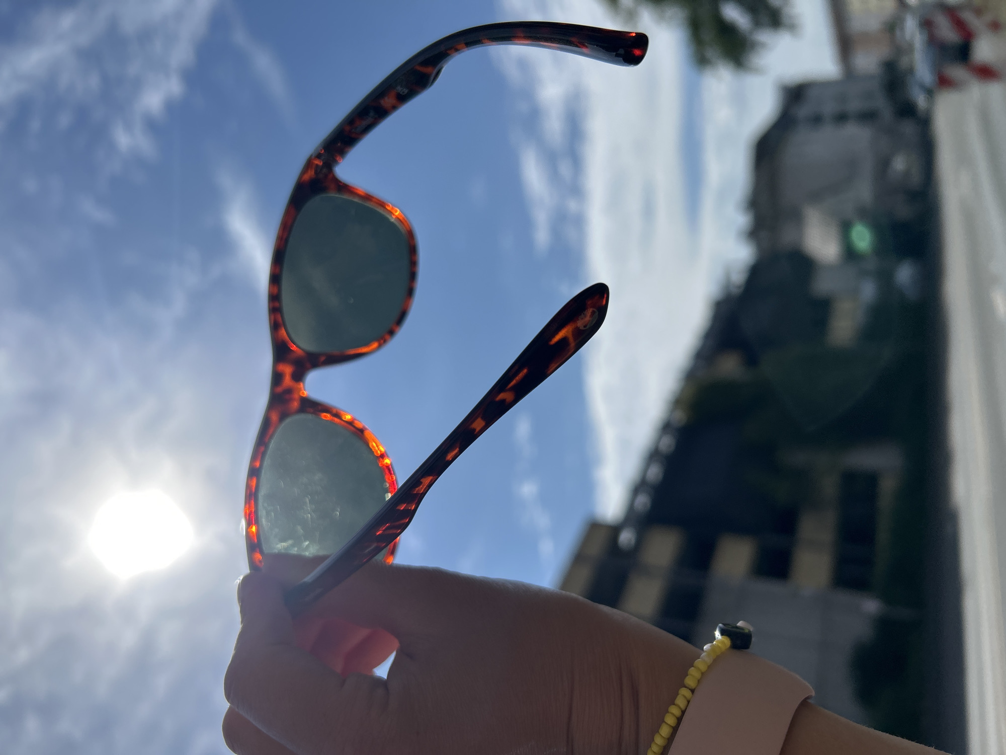 A person's hand holds a pair of tortoise-shell pattern sunglasses up to the bright sun against a blue sky with wispy clouds. The sunglasses are angled so the lenses are visible, revealing a greenish tint. The hand is wearing a light pink watchband with a yellow beaded bracelet. In the background, out of focus, is a multi-story building with dark windows, and further beyond, other buildings and trees are visible, suggesting an urban environment. The bright sun obscures details in the sky and creates a lens flare. The scene appears to be outdoors in a city.