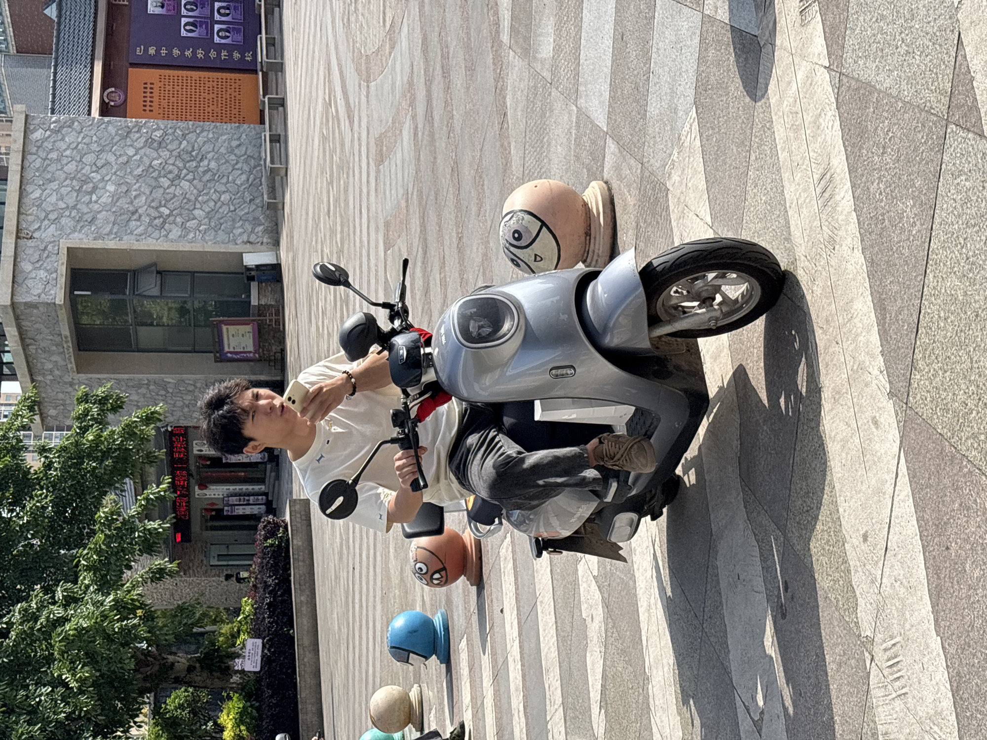 This image depicts an outdoor urban scene on a clear, sunny day, likely in a pedestrian area in Jiangjin, China. A young man is seated on a silver-grey electric scooter or moped, paused on a paved surface. He has dark hair, is wearing a light-colored (white/cream) short-sleeved t-shirt, and dark trousers, with a bracelet on his left wrist. He is holding a light-colored smartphone in his left hand and appears to be focused on its screen, while his right hand rests on the scooter's handlebar.
The ground is paved with a pattern of square and rectangular tiles in shades of grey and beige, with long, distinct shadows cast, indicating bright sunlight. In the background, there are buildings. To the left, a building features a grey, stone-like facade and a partially open window. Higher up and further back, another building displays a large, dark purple sign with visible white Chinese text: "巴南中学安全生产学校" (Banan Middle School Safety Production School) and "巴南区教育委员会" (Banan District Education Committee). Below this is an orange, textured section. Green foliage from trees and shrubs is visible on the left side of the frame.
A distinctive feature of the setting is a series of colorful, spherical bollards or decorative elements lining the paved area. These spheres are painted with cartoonish faces, adding a playful element to the environment. One light brown bollard close to the scooter features large eyes and a downturned mouth, while a blue and an orange one are also visible further down. The scene captures a casual moment in a public space, possibly a plaza or a wide sidewalk.