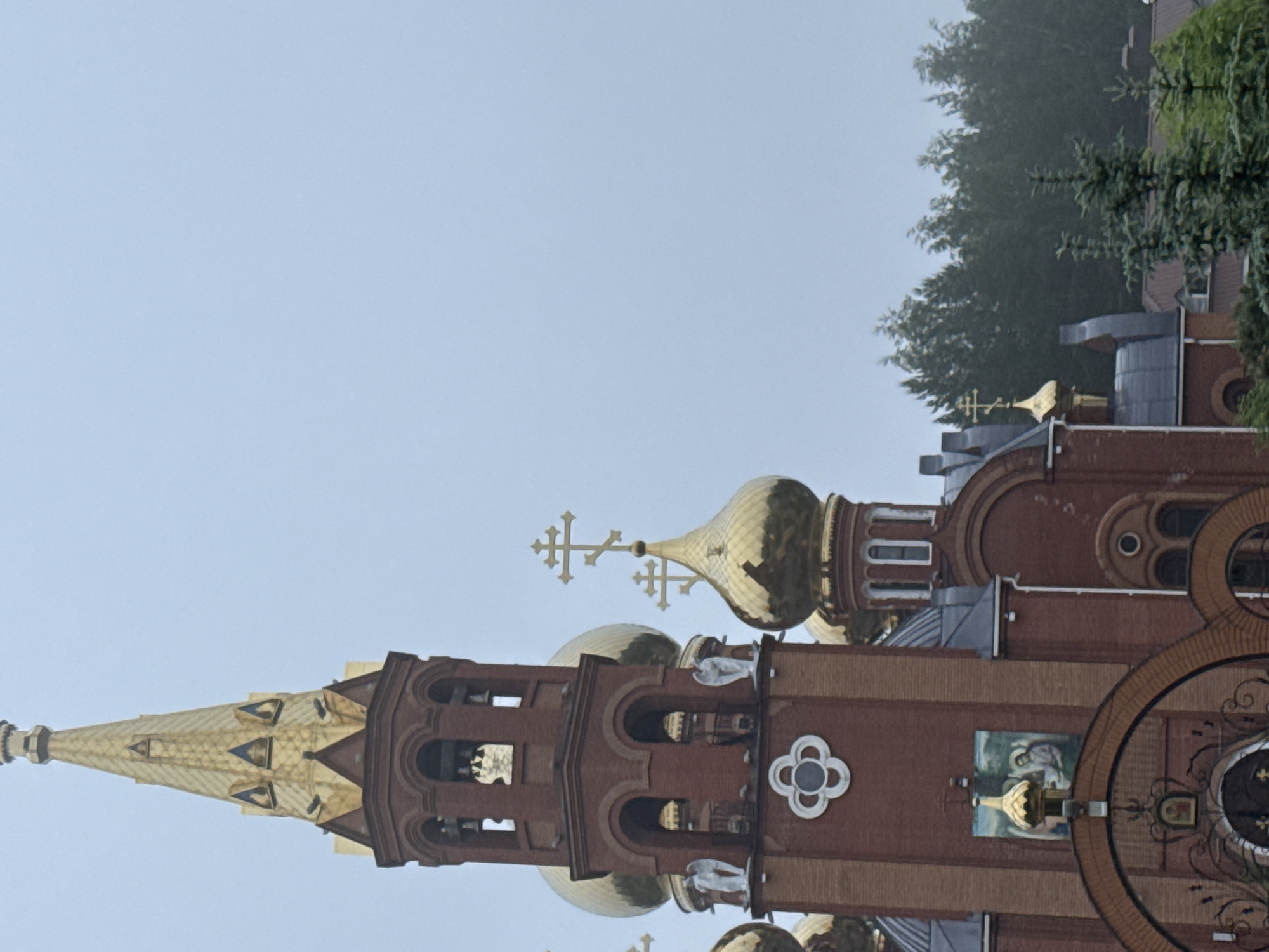 The media file captures a close-up view of an ornate Eastern Orthodox church, likely located in Chaikovsky, Russia, as indicated by the context. The church is constructed from reddish-brown brick and features multiple prominent golden onion domes, each topped with a distinct Orthodox cross. A tall, golden spire, intricately designed, also rises majestically towards the left side of the frame.
The architectural details include arched windows and decorative elements, such as a white quatrefoil design, embedded in the brickwork. On one of the exterior walls, a fresco or mosaic depicting a religious figure, possibly Jesus or a saint, is visible. Green trees and foliage are present in the background on the right side, suggesting the church is set within a natural or landscaped environment.
The scene appears to be taking place during the daytime, under an overcast or cloudy sky, which provides soft, diffused lighting. No direct activity is depicted; the image is a static architectural photograph focusing on the grandeur and details of the church. No discernible text is visible on the building.