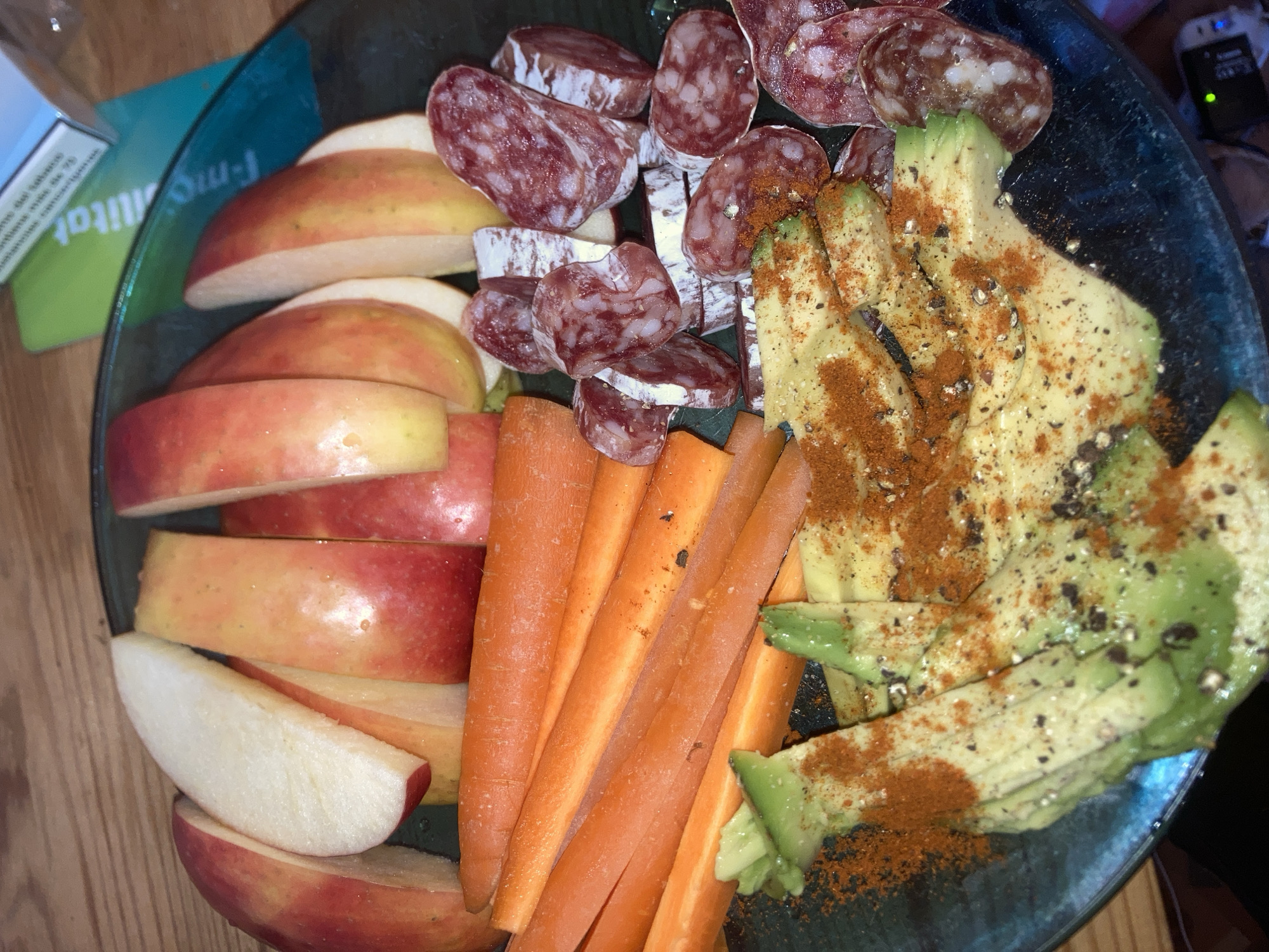 The scene depicts a close-up view of a meal or snack presented on a dark, possibly translucent blue-green glass plate. The scene is located in the city of Barcelona, Spain.
On the plate, arranged from left to right:
* Several slices of red and yellow apples, some with skin and some peeled, are positioned along the left and top edge of the plate.
* Below the apple slices, a cluster of raw, whole carrots is visible.
* Further down on the left, sliced avocado pieces are spread, heavily seasoned with ground black pepper and a reddish-orange spice, likely paprika or chili powder.
* On the right side of the plate, numerous slices of cured sausage or salami are displayed, exhibiting characteristic marbling and a white casing or bloom on some pieces.
The plate rests on a wooden surface, visible in the background, particularly in the upper left and upper right portions of the image. In the upper right background, a light blue and green rectangular object, consistent with a cigarette pack, is partially visible. Text fragments on its green section include "f-mobi litat," and text on the blue section appears to be in French, stating "fume du tabac" and the number "25." Below the plate on the bottom right, several electronic devices are partially visible, one featuring a distinct green indicator light. No individuals are present in the image.