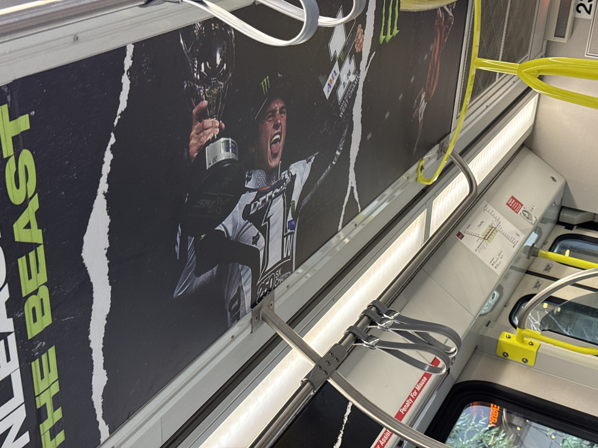 The image shows the interior of a public transit vehicle, likely a bus or train, in Calgary, Canada. The ceiling of the vehicle is covered with a large, dark advertisement featuring a distressed, ripped texture. Prominently displayed in bright green lettering are the words "THE BEAST". Below this, a photograph shows a person wearing a motocross helmet and gear, with their mouth open in a yell or laugh. The person is holding a trophy and is likely a motocross athlete. The photograph appears to be a close-up, with the rider's face angled towards the viewer. The advertisement also includes logos for "AMA" and "Monster Energy".
Inside the vehicle, there are metal handrails and straps for passengers to hold onto, with some straps hanging down. There are also yellow grab poles and fluorescent lighting panels along the ceiling. On the wall below the advertisement, there are various signs and notices, including a red sign that reads "Penalty For Misuse" and what appears to be a route map or diagram. The windows of the vehicle show a blurred outdoor scene, suggesting movement. The overall impression is of an advertisement campaign on public transport, common in urban environments like Calgary.