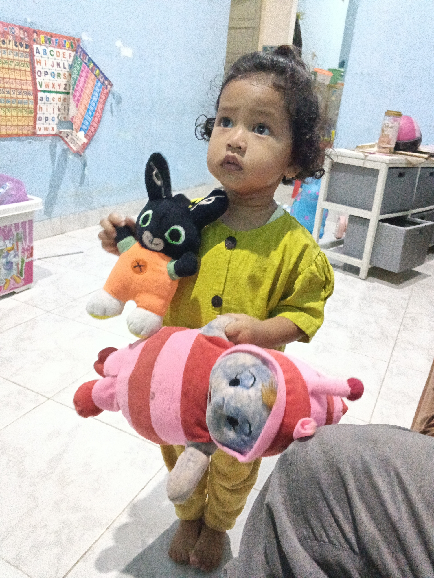 A young girl with curly dark hair stands in a room, holding two stuffed animals. The girl is wearing a yellow shirt and yellow pants. She is looking upwards and to her right. In her right hand, she holds a black bunny-like stuffed animal with green eyes and an orange outfit. Below the bunny, she holds a larger stuffed animal with pink and red stripes and a pink hood. The room has a light blue wall with an alphabet chart and a number chart. A white dresser with grey drawers and a pink object on top is to the right of the girl. To the left of the girl, there is a white plastic bin with a pink lid and a sticker of a cartoon character. The floor is tiled in white. The scene appears to be a residential interior.