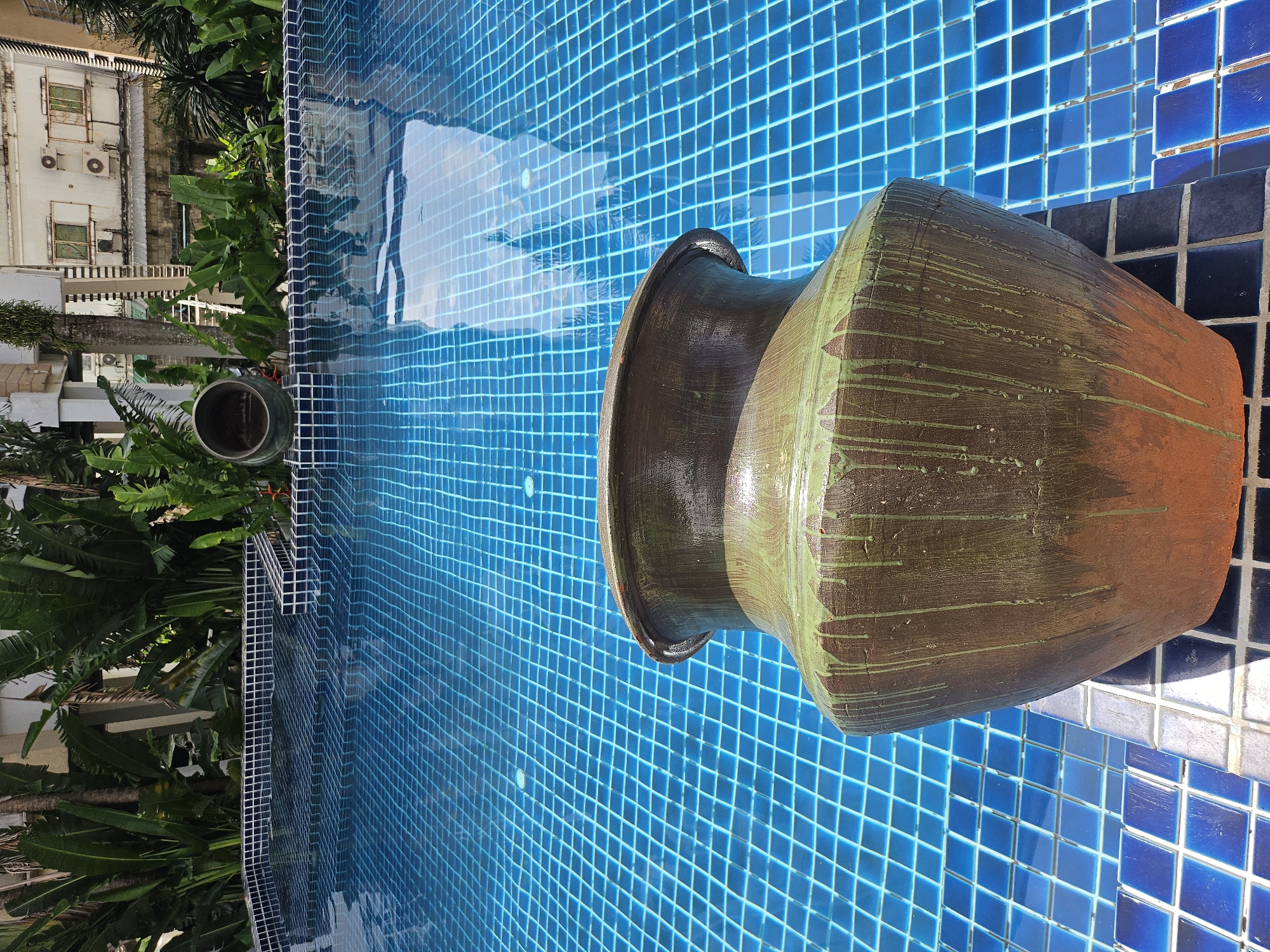 A large, brown, terra cotta pot with green drippings sits on the tiled edge of a bright blue swimming pool. The pool is tiled with small, square, dark blue tiles. In the background, lush green tropical foliage is visible, along with parts of a multi-story building. The sky and surrounding vegetation are reflected in the clear water of the pool. The scene appears to be a residential or hotel setting in Kathu, Thailand. There are no people visible in the image.