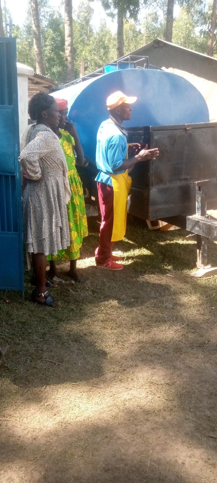 The image shows three people standing in front of a blue water tank on a dirt lot in Kivaywa, Kenya. The location is confirmed using the provided latitude and longitude coordinates. The people are all dressed in casual clothing. The person on the left is wearing a white dress, the person in the middle is wearing a green and yellow dress, and the person on the right is wearing a blue shirt, yellow apron, and maroon pants. The man on the right is pointing his right hand at something behind the camera. The ground is covered in dry grass and dirt. There is a blue door in the background on the left side of the image. There is a large blue water tank in the background that is on a metal frame. The tank is partially covered by a metal roof. There is a building in the background on the right side of the image. The scene is bright and sunny. The sky is clear. There are trees in the background.