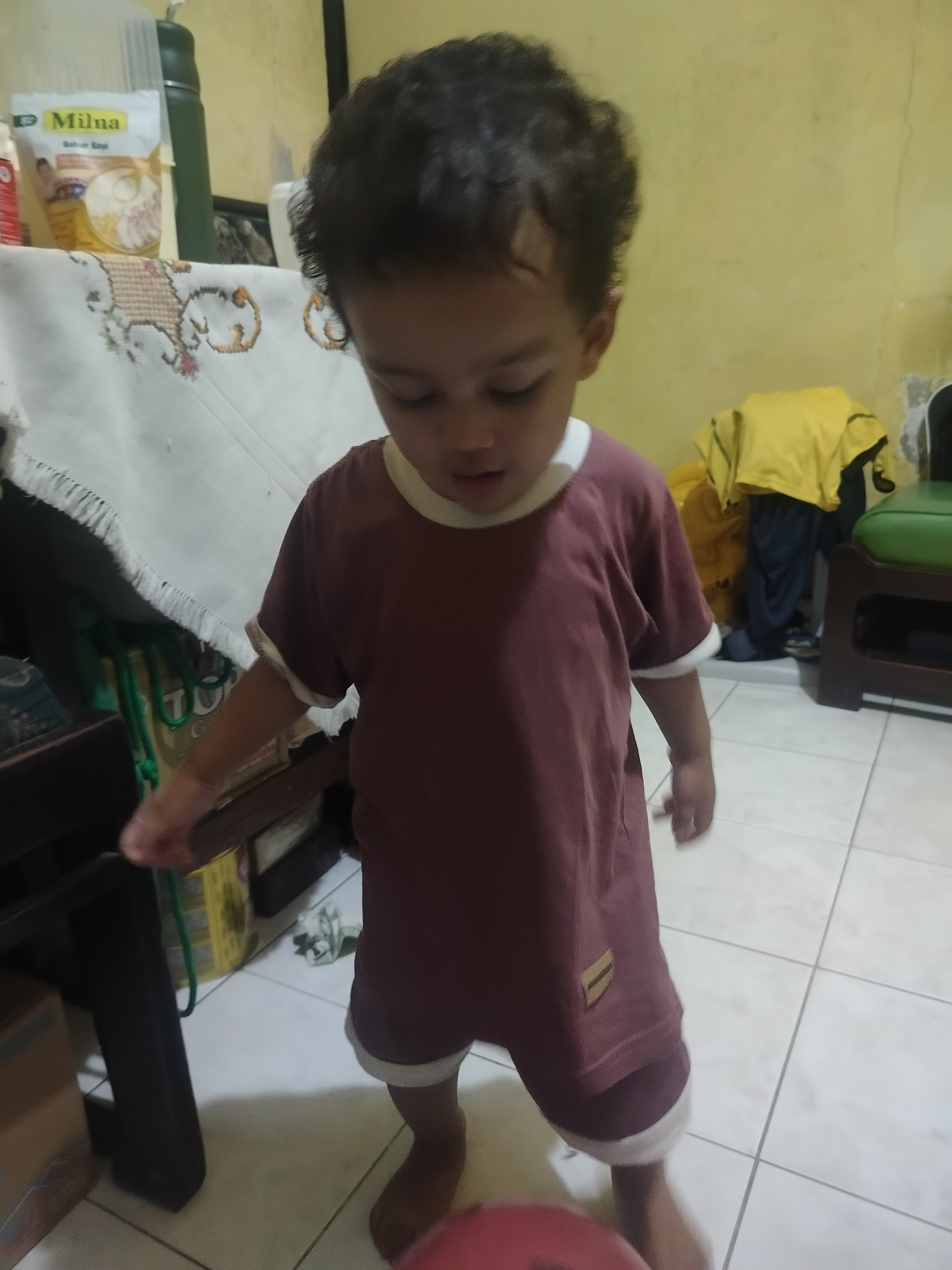 A young child, possibly a toddler, stands in an indoor setting. The child has dark, curly hair and is wearing a maroon t-shirt with white trim on the sleeves and collar, and matching maroon shorts with white trim at the hem. The child is looking down and to the right, with their left hand extended downwards and slightly forward. The floor appears to be tiled in a light color, with grout lines visible. In the background, there is a yellow wall, a table covered with a white cloth that has an embroidered design, and some items on shelves or in the background, including a green thermos, a white packet labeled "Milna", and some clothing items, including a yellow shirt and blue pants. There is also a dark wooden table or cabinet to the left of the child. The overall lighting suggests an indoor environment, possibly a home. The scene is consistent with a domestic interior in Surabaya, Indonesia.