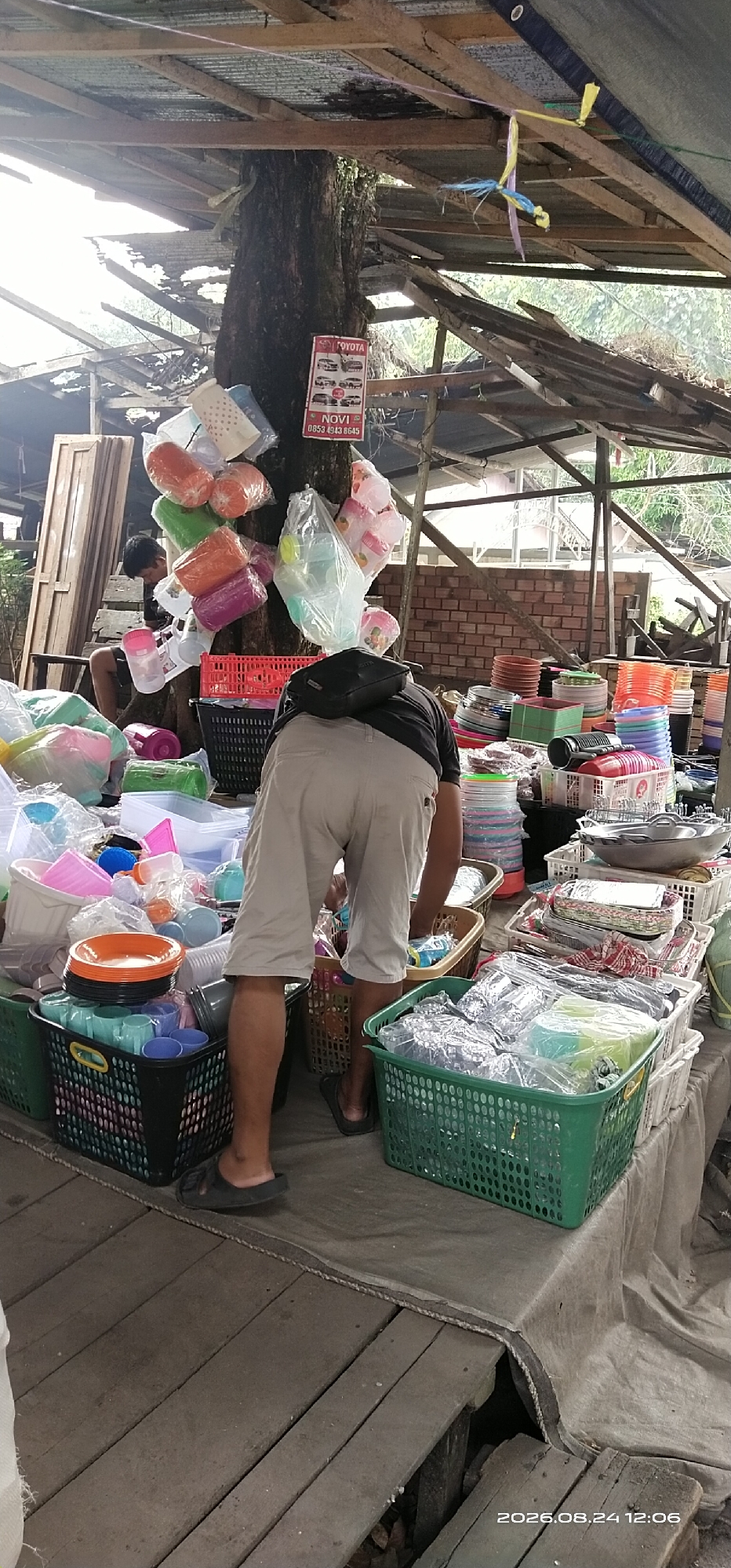 The image depicts an outdoor market scene, likely in Tamiang Layang, Indonesia, characterized by a rustic wooden structure providing shelter. A large tree trunk is prominent, serving as a central support for the overhead roofing and also as a display for various plastic household items, such as containers and cups, bundled in plastic bags.
In the foreground, a person wearing light-colored shorts and dark sandals is bent over, interacting with goods displayed on a table. This table is covered with a grey tarp and laden with an assortment of plastic storage containers, bowls, and plates, stacked in baskets and on the surface. The containers vary in color, including green, pink, blue, and orange. To the left, another person, a young male wearing a dark t-shirt and shorts, is seated, also appearing to be involved in the market activity.
The environment suggests a modest setup, with wooden planks forming the floor and the rickety roofing made of corrugated metal and timber. In the background, a brick wall is visible, and beyond that, dense foliage indicates a natural, possibly rural, setting. A sign displaying images of Toyota vehicles and contact information for "NOVI" is attached to the tree. The overall impression is of a functional, albeit somewhat makeshift, marketplace.