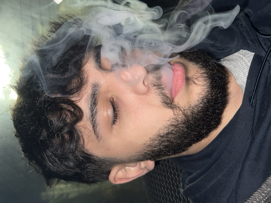 A male individual is depicted in a close-up, chest-up shot, located in Al-Muhajerin Municipality, Syria. The subject has dark, curly hair and a full, dark beard. His eyes are closed. A plume of white-grey smoke is visibly exhaling from his mouth, partially obscuring the right side of his face and circulating around his head. He is wearing a dark, possibly navy, hooded sweatshirt over a lighter grey garment. The background is dark and out of focus, revealing a dark, textured surface to the left, consistent with an interior or vehicle environment.